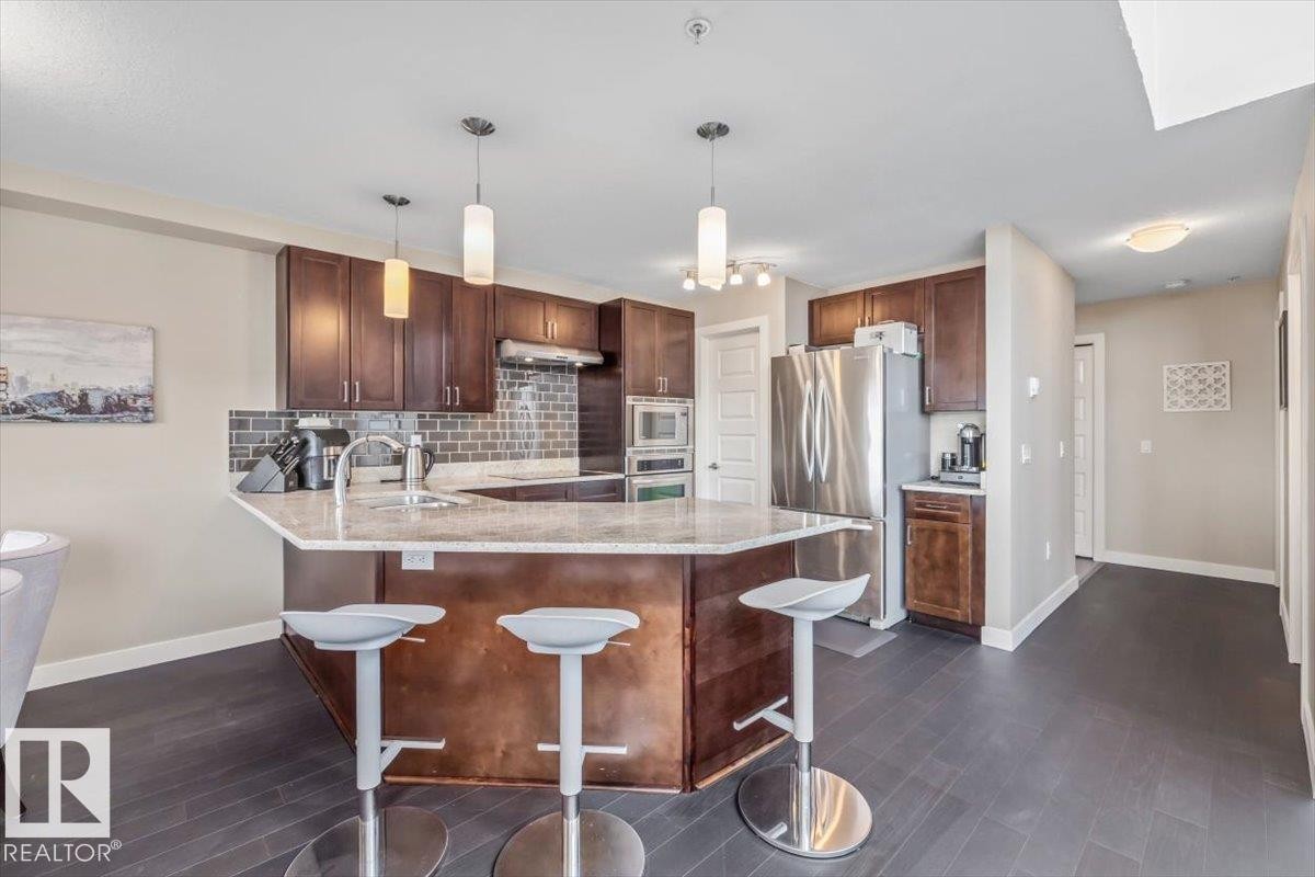 Kitchen featuring dark wood cabinetry, a granite-finish island with seating, stainless steel appliances, a tile backsplash, and recessed and pendant lighting - 513 1230 Windermere Way, Edmonton, AB