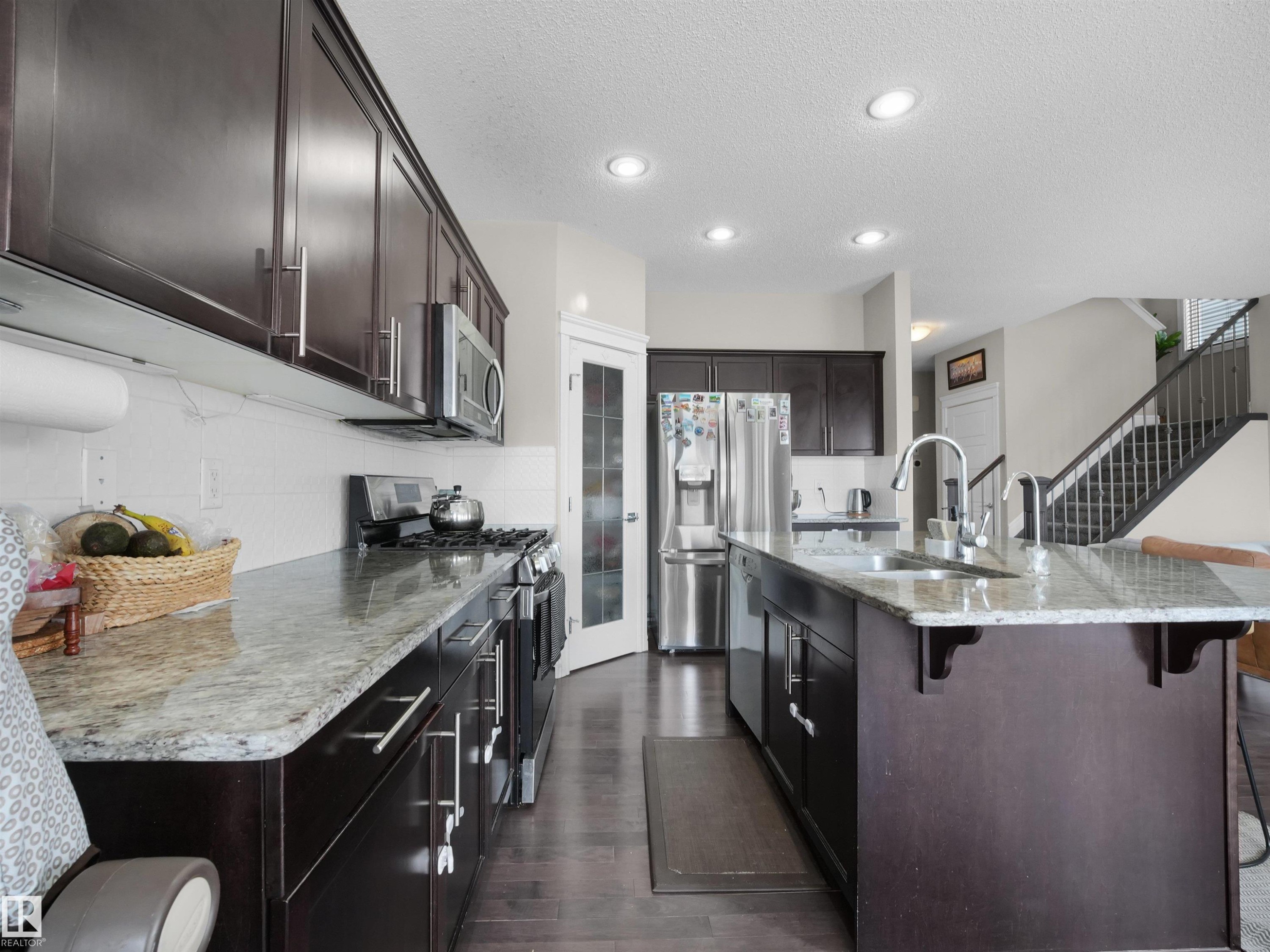 Kitchen featuring dark wood cabinetry, light-toned countertops, stainless steel appliances, a center island with sink, and recessed lighting - 3247 15 Avenue Nw, Edmonton, AB