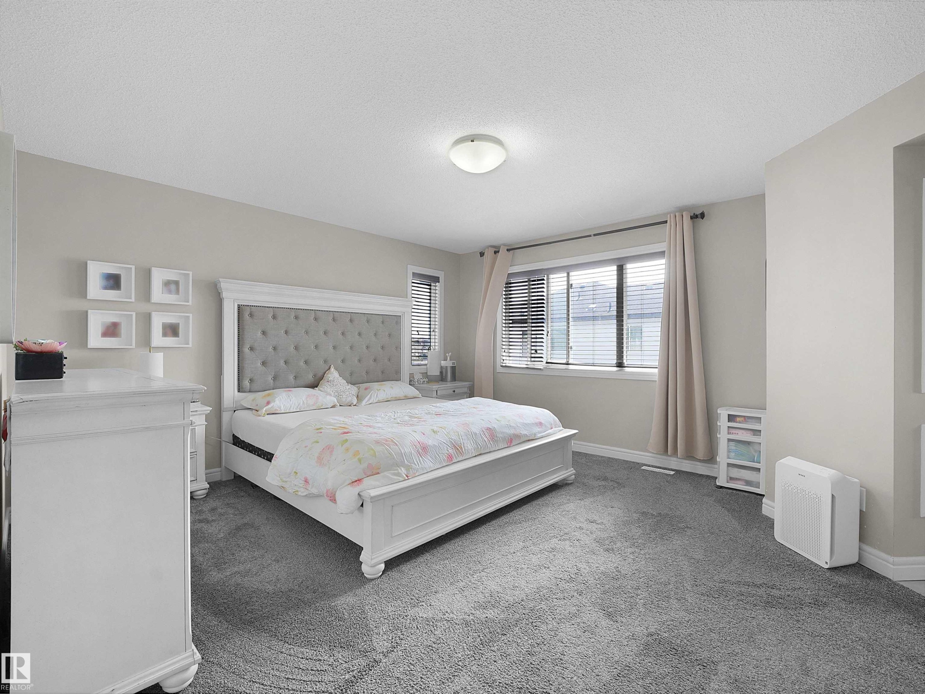 Spacious bedroom featuring neutral wall tones, grey carpeting, a large window with blinds and drapes, and a flush-mount ceiling light - 3247 15 Avenue Nw, Edmonton, AB
