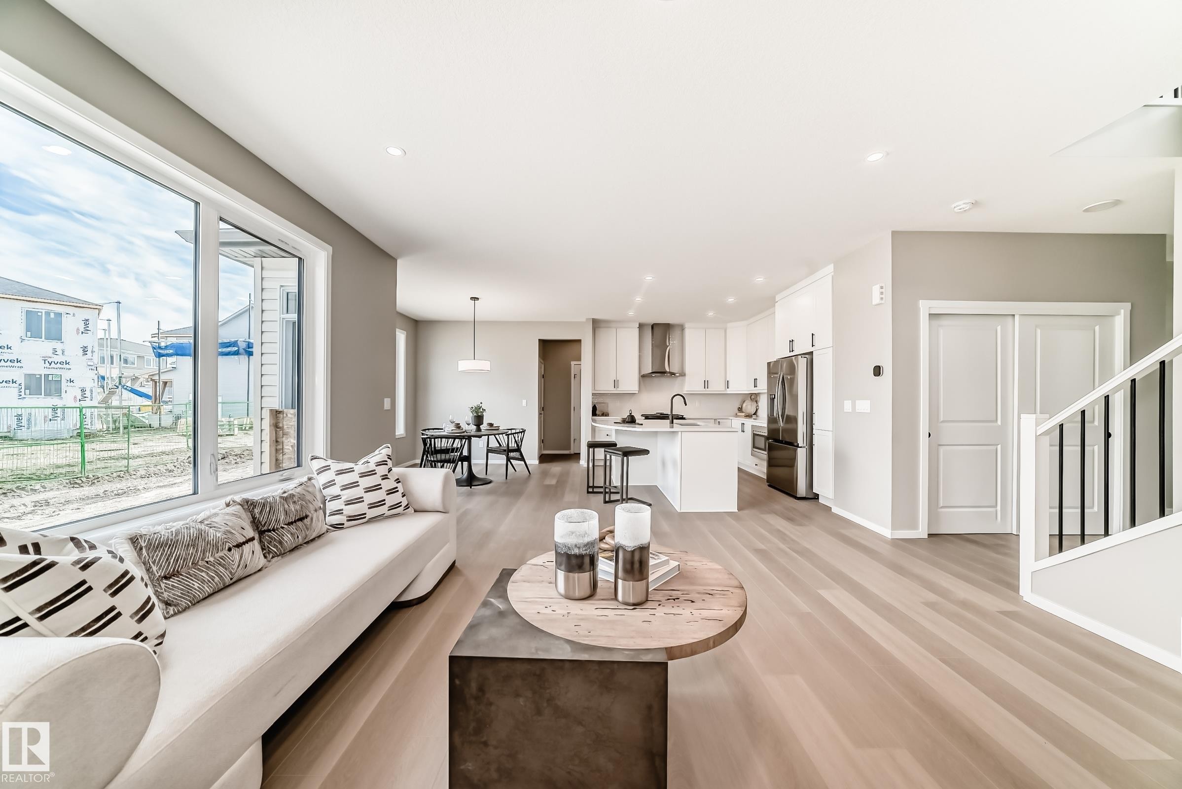 Expansive open-concept living area featuring large windows, light wood-finish flooring, a contemporary kitchen with white cabinetry and stainless steel appliances, and a dining area with modern pendant lighting - 1603 206 Street, Edmonton, AB - Indoor Photo Showing Living Room