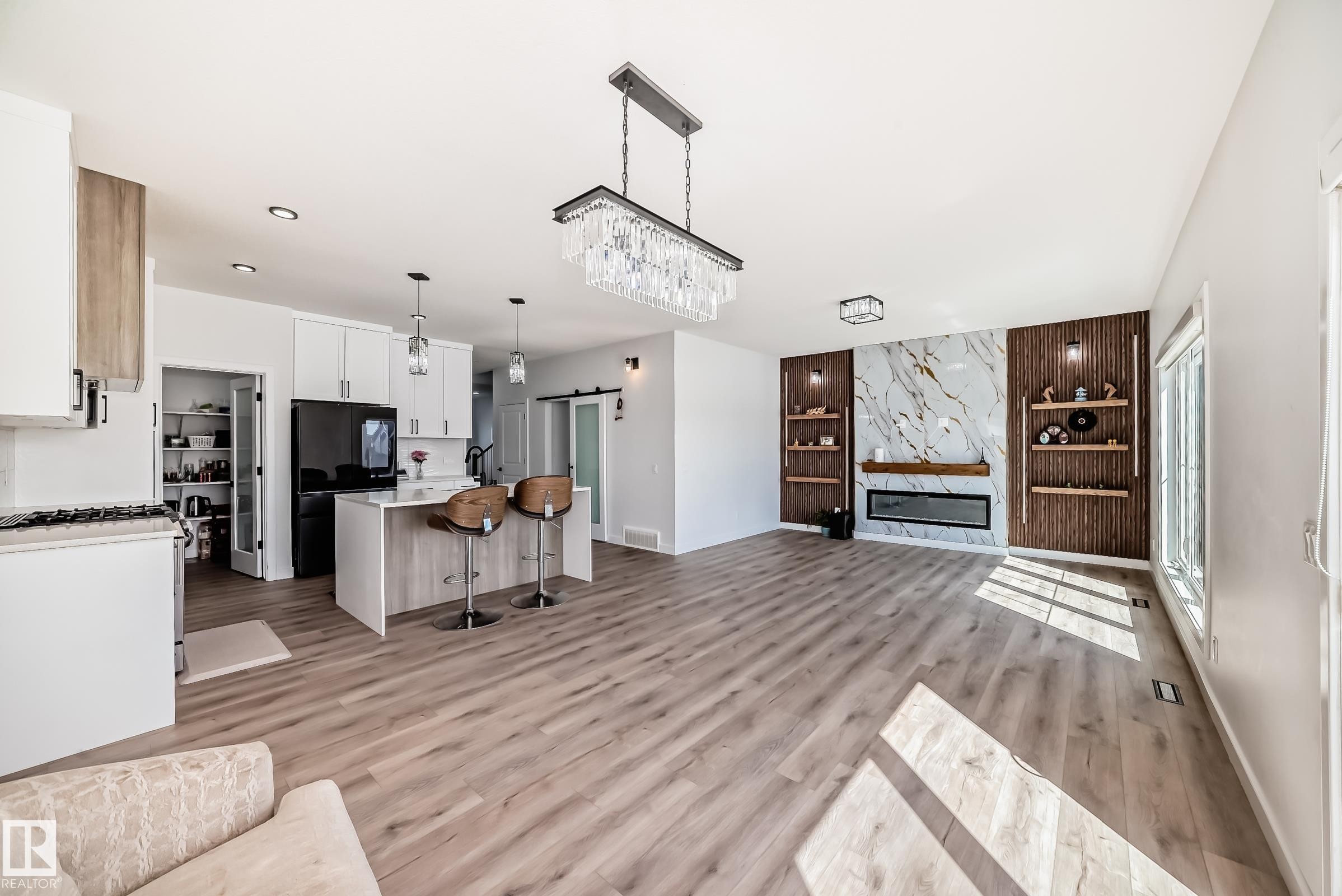 Open-concept living space featuring wood-finish flooring, a modern linear fireplace with a marble-effect surround, built-in shelving, a kitchen island with pendant lighting, and white cabinetry - 3247 Chernowski Way, Edmonton, AB - Indoor