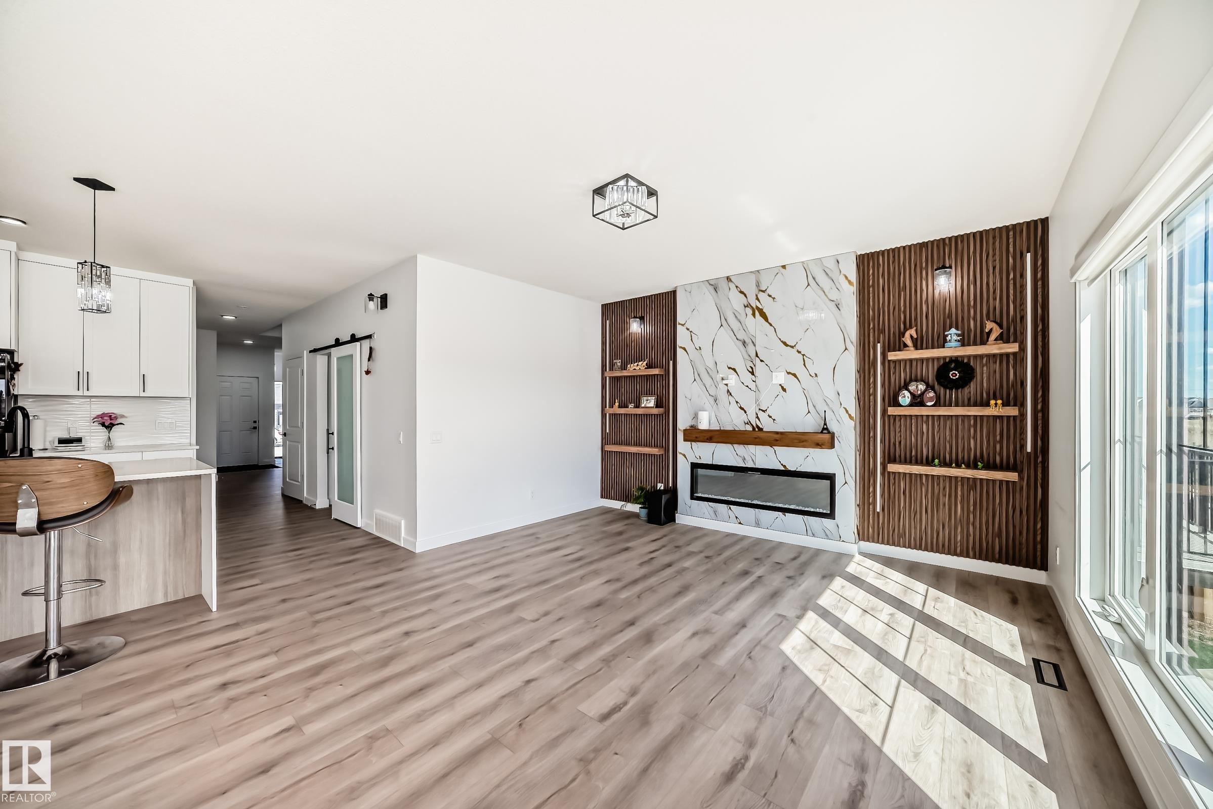 Bright open-concept living area featuring wood-finish flooring, a contemporary linear fireplace set into a large-format marble-look accent wall, and integrated shelving with vertical slat paneling - 3247 Chernowski Way, Edmonton, AB - Indoor