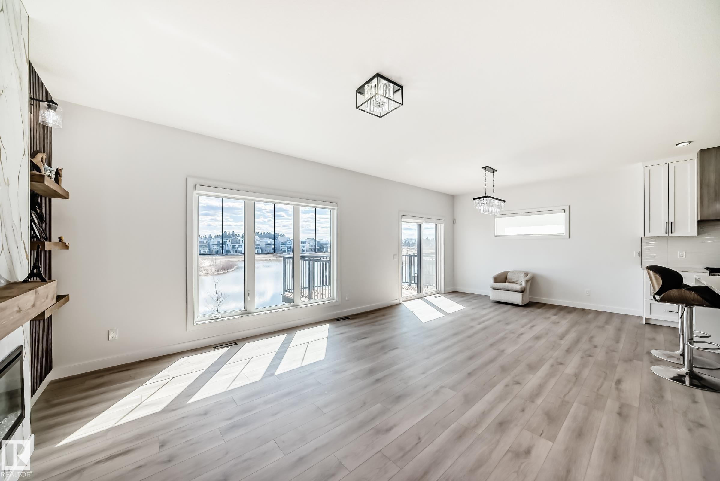 Spacious open-concept room featuring light wood-finish flooring, white walls, and a large multi-pane window overlooking a body of water - 3247 Chernowski Way, Edmonton, AB - Indoor With Fireplace