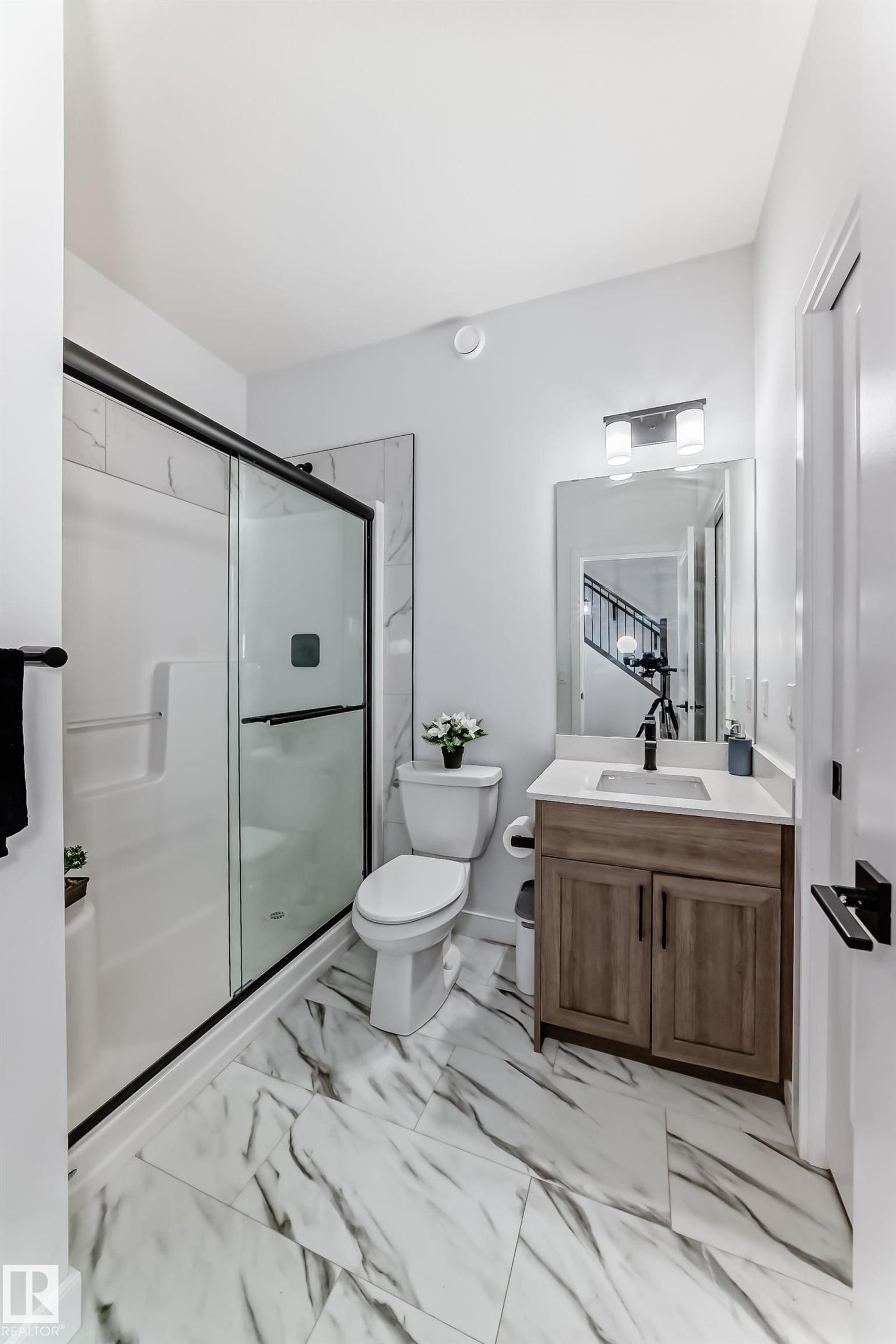 Modern bathroom featuring large format marble-look tile flooring, a glass-enclosed shower with black framing, a light wood-finish vanity with a white countertop, and a contemporary wall-mounted light fixture - 3247 Chernowski Way, Edmonton, AB - Indoor Photo Showing Bathroom