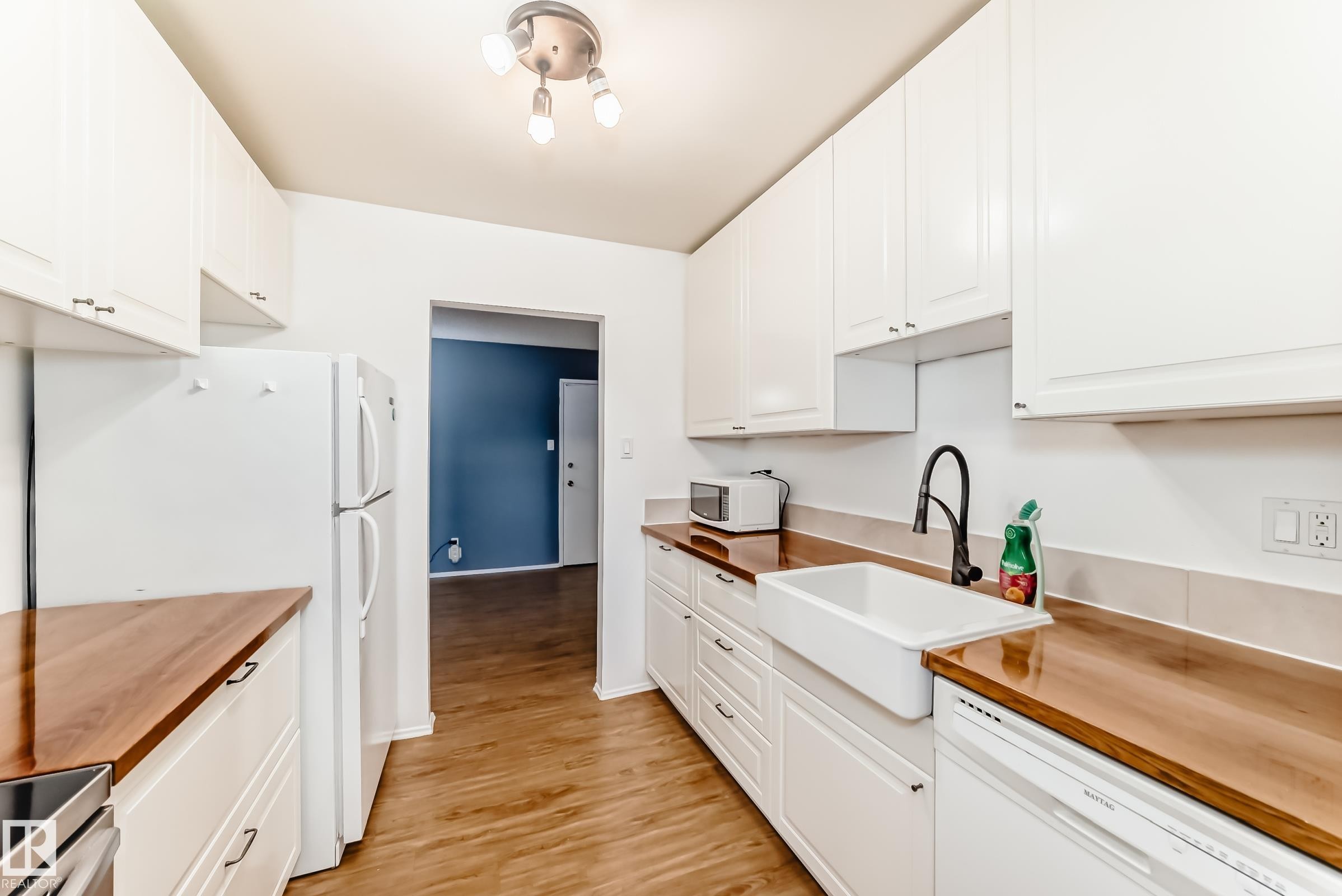 Kitchen featuring white cabinetry, wood-finish countertops, wood-finish flooring, a farmhouse-style sink with a matte black faucet, and white appliances - 423 5125 Riverbend Road, Edmonton, AB - Indoor Photo Showing Kitchen