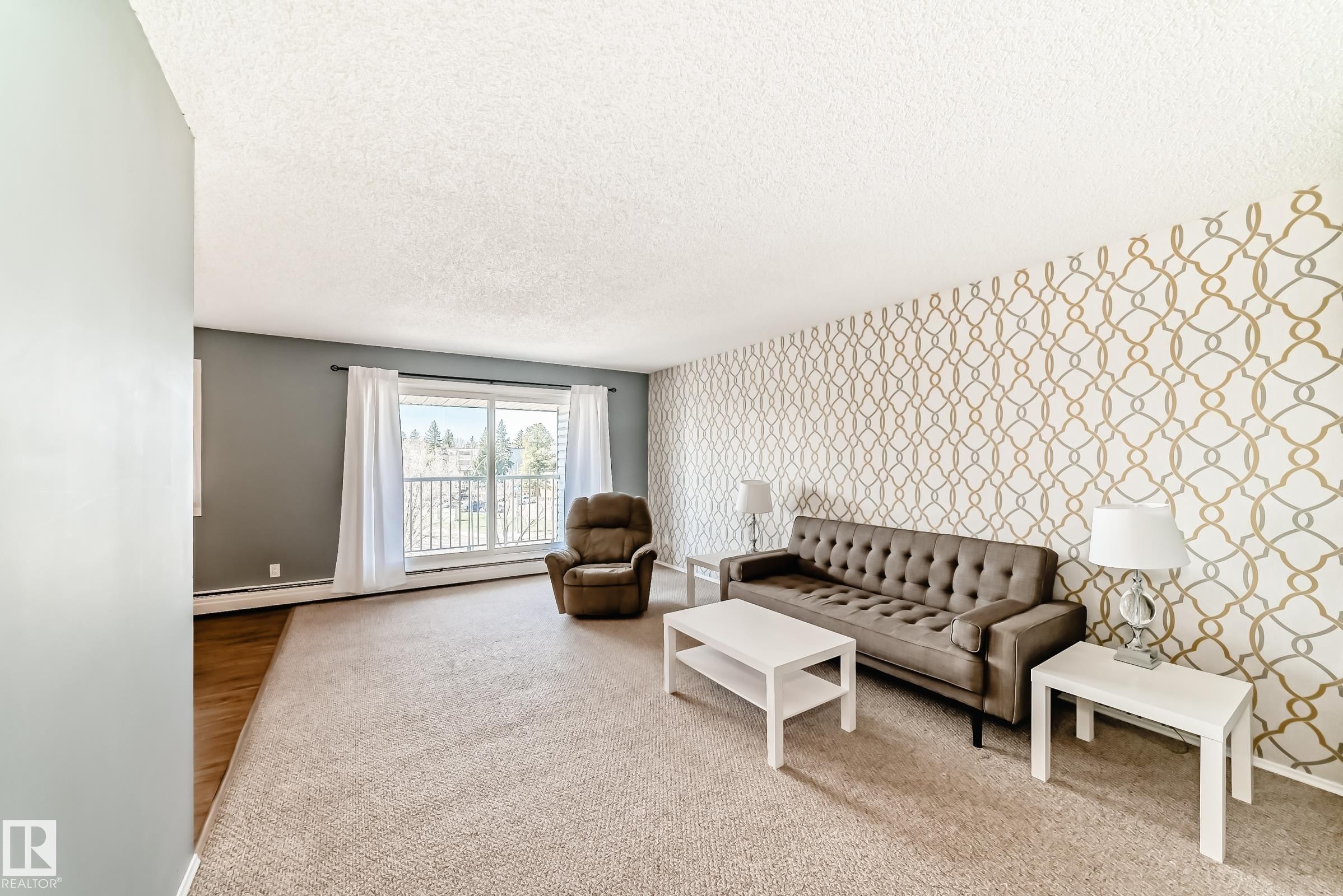 Spacious living area featuring a large window with white vertical blinds, patterned accent wall, and textured carpeting - 423 5125 Riverbend Road, Edmonton, AB - Indoor