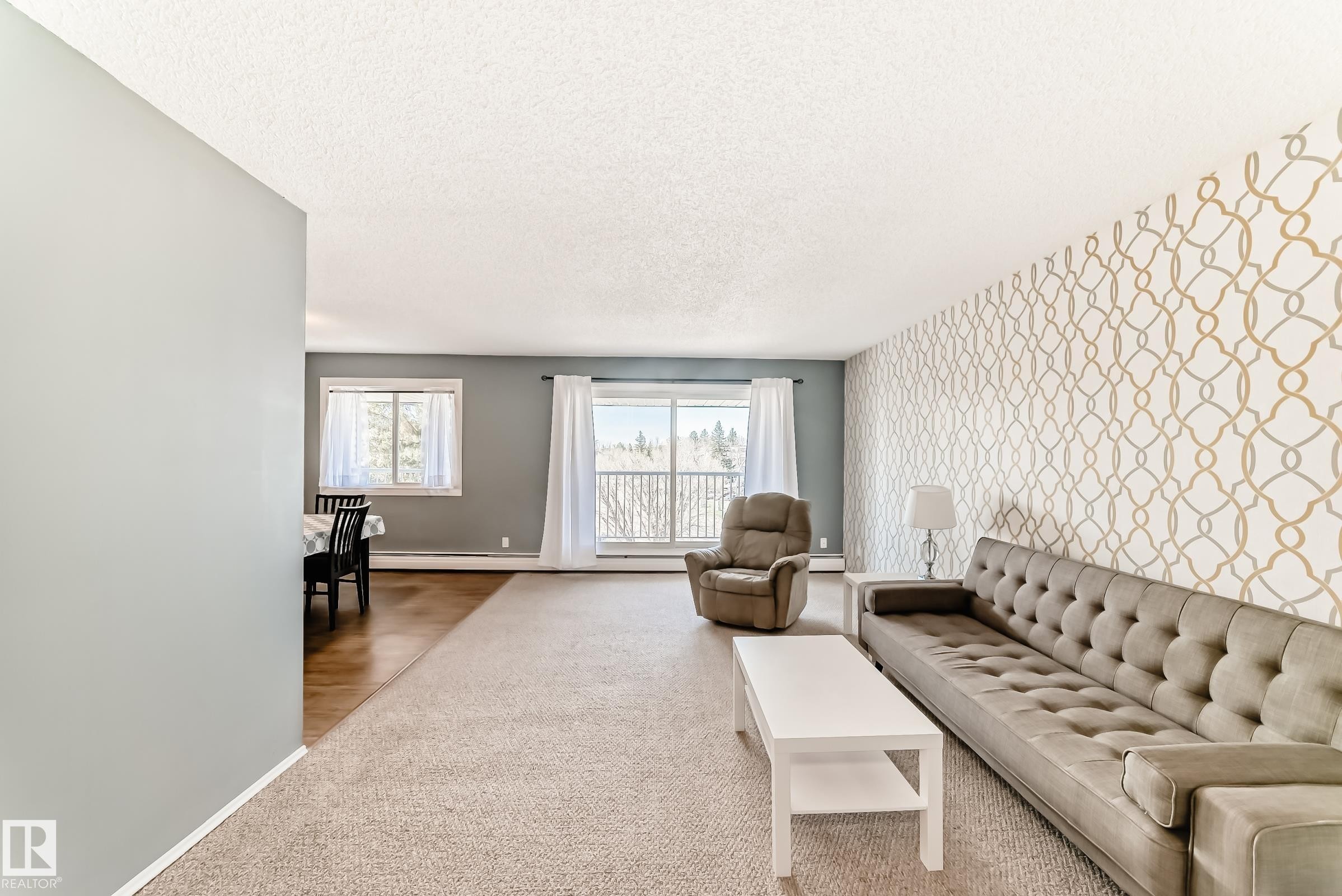 Spacious living area featuring patterned accent wall, light neutral carpeting, and a large window - 423 5125 Riverbend Road, Edmonton, AB - Indoor