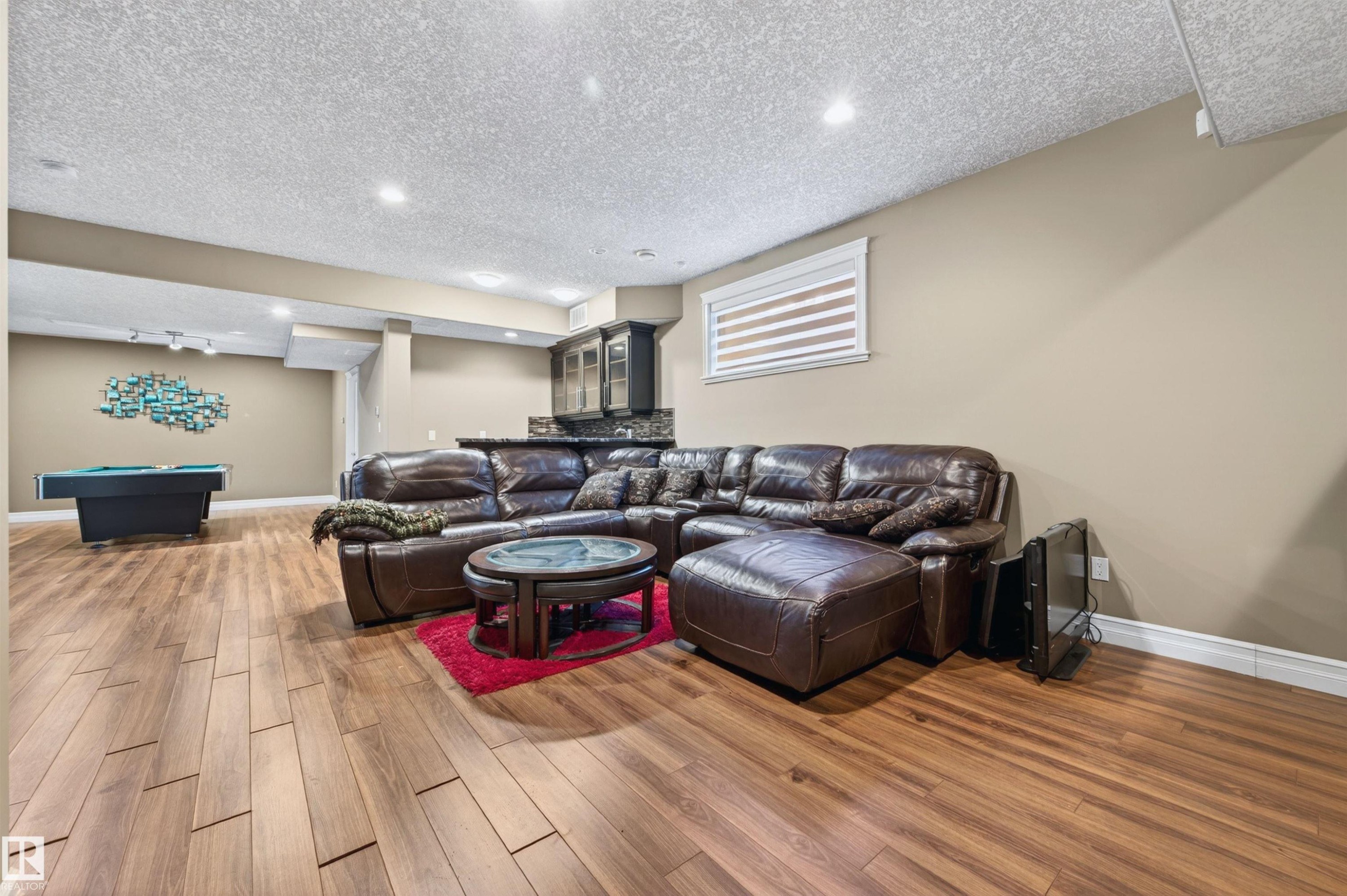 Spacious open-concept lower level with wood-finish flooring, recessed lighting, and a built-in wet bar featuring dark cabinetry and a stone-finish countertop - 1254 Adamson Drive, Edmonton, AB - Indoor