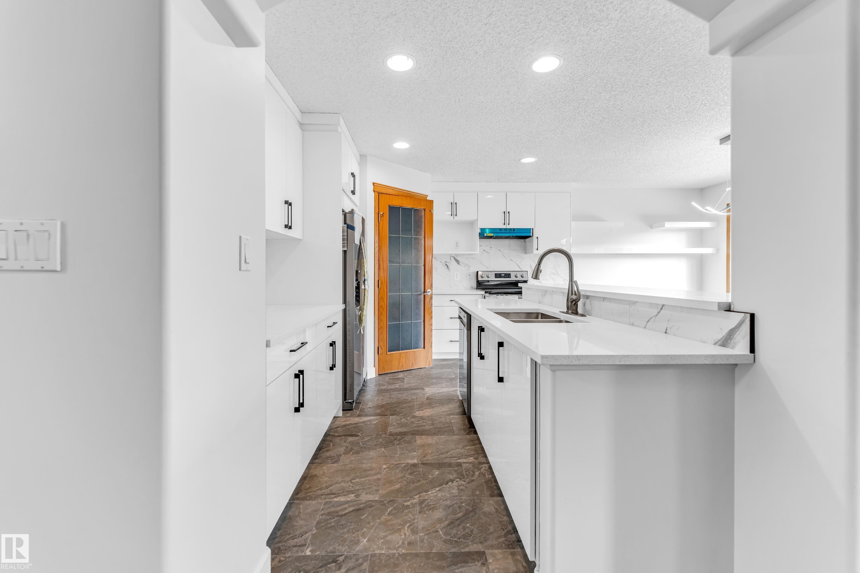 Contemporary kitchen featuring white cabinetry, a large kitchen island with an undermount sink and gooseneck faucet, light-toned countertops with a matching backsplash, and dark tile flooring - 10621 175A Avenue, Edmonton, AB
