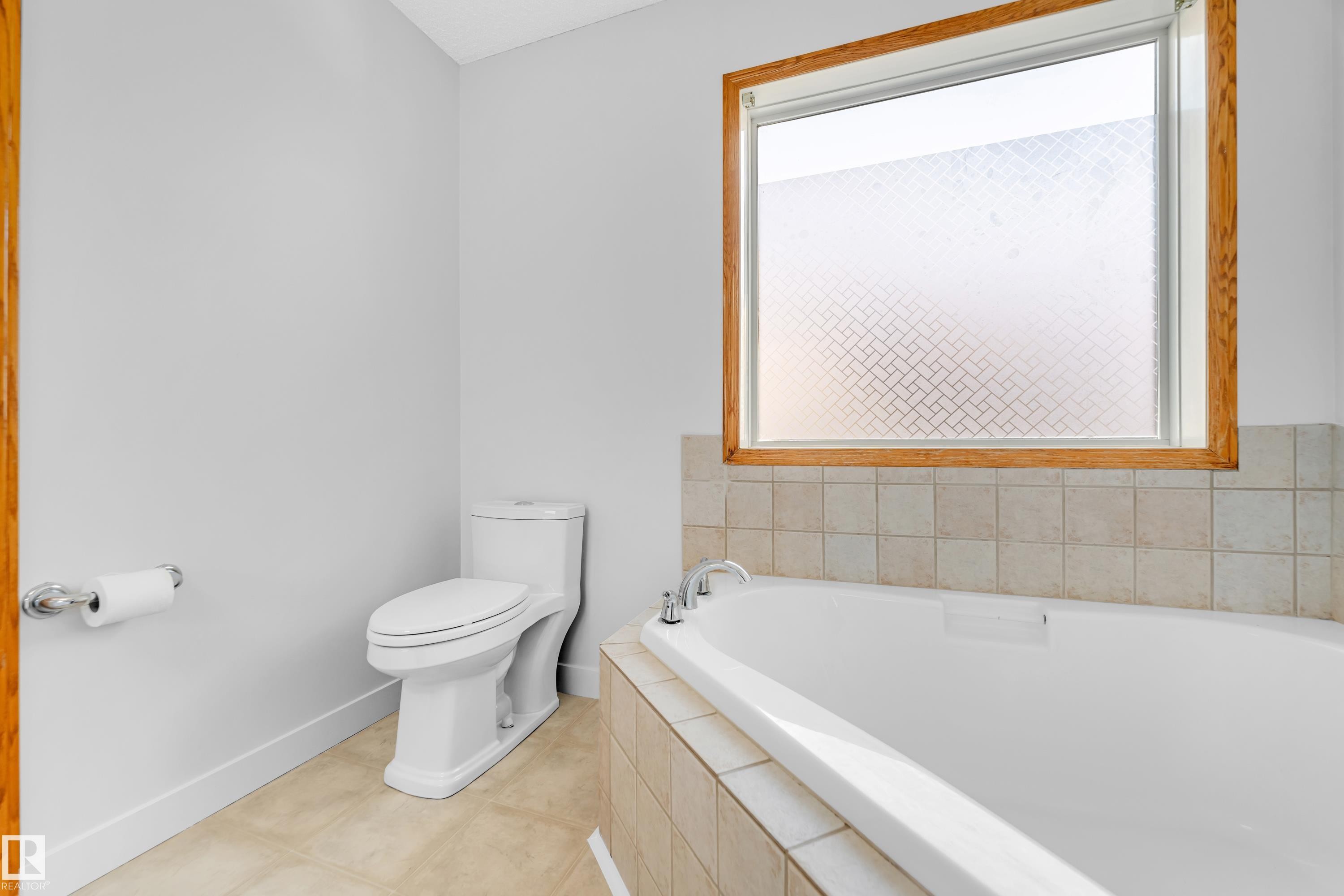 Bathroom featuring a built-in soaking tub with tile surround, a large window with wood trim, and light-toned tile flooring - 10621 175A Avenue, Edmonton, AB