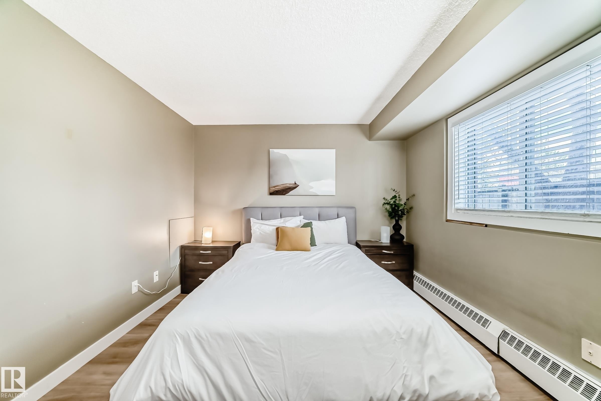 Bedroom featuring a large window with blinds, wood-finish flooring, and neutral wall tones - 101 10827 85 Avenue Nw, Edmonton, AB - Indoor Photo Showing Bedroom