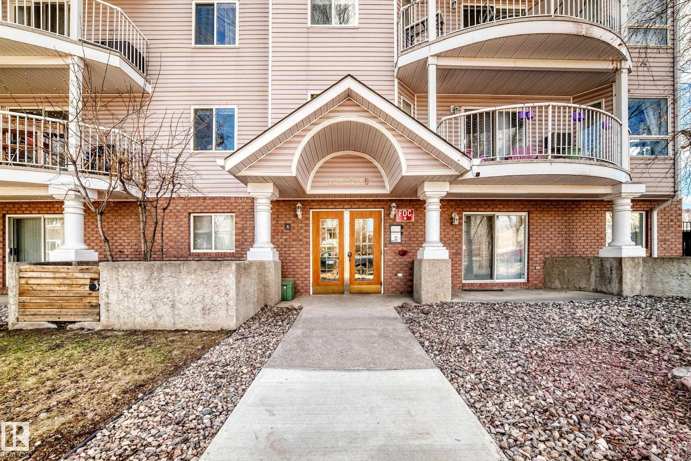 Building exterior featuring brick and siding finishes, a covered entrance with double doors, curved balconies with white railings, and a concrete pathway - 101 10827 85 Avenue Nw, Edmonton, AB - Outdoor
