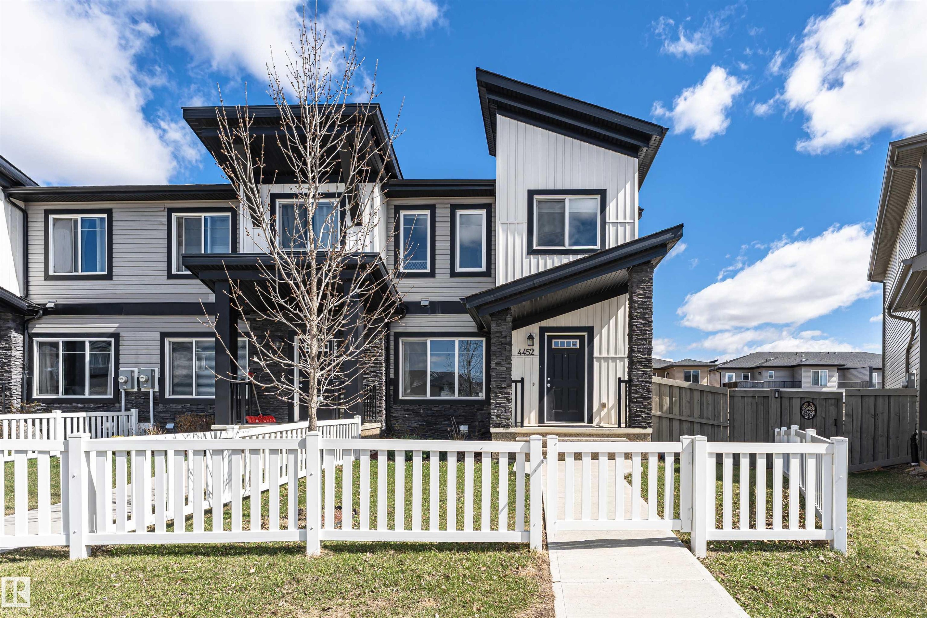 Modern townhome exterior featuring an angular roofline, board and batten siding, stone facade accents, a black entry door, and a white picket fence - 4452 Annett Common, Edmonton, AB