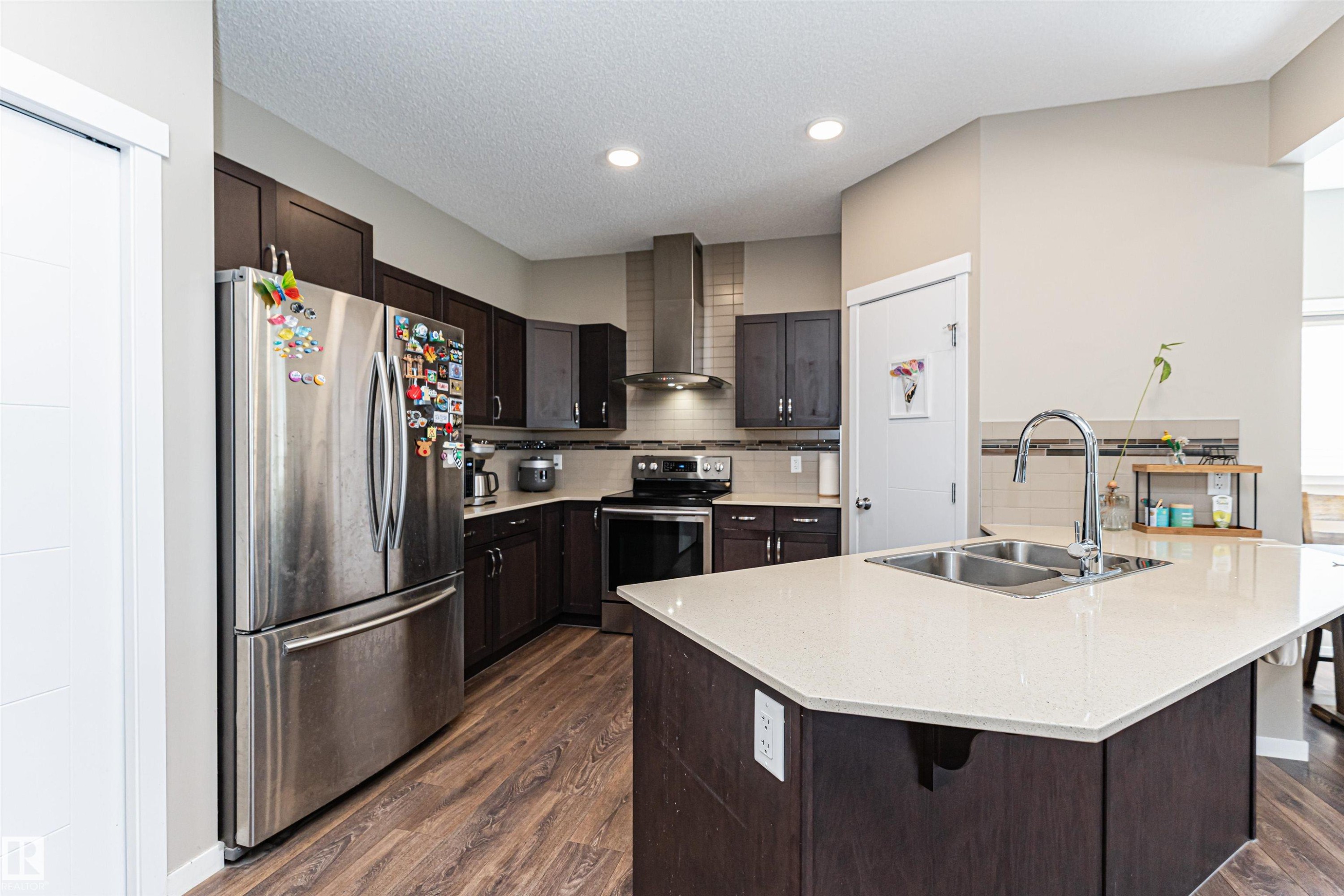 Kitchen featuring a substantial island with a double basin sink and a chrome faucet, dark wood-finish cabinetry, stainless steel appliances, recessed lighting, and wood-finish flooring - 4452 Annett Common, Edmonton, AB