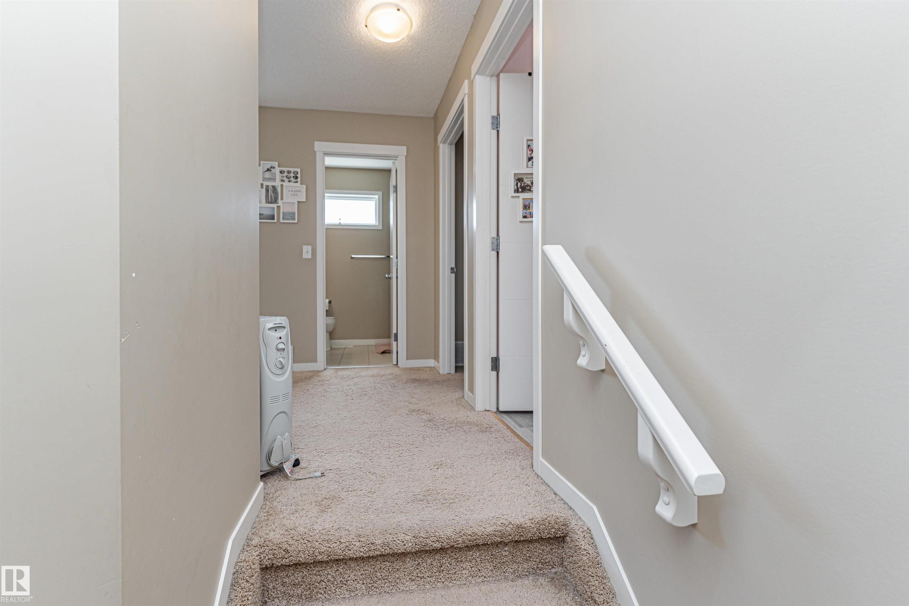 Carpeted hallway with light neutral wall paint and white trim, featuring a flush mount ceiling light, a built-in white handrail, and two interior doorways - 4452 Annett Common, Edmonton, AB