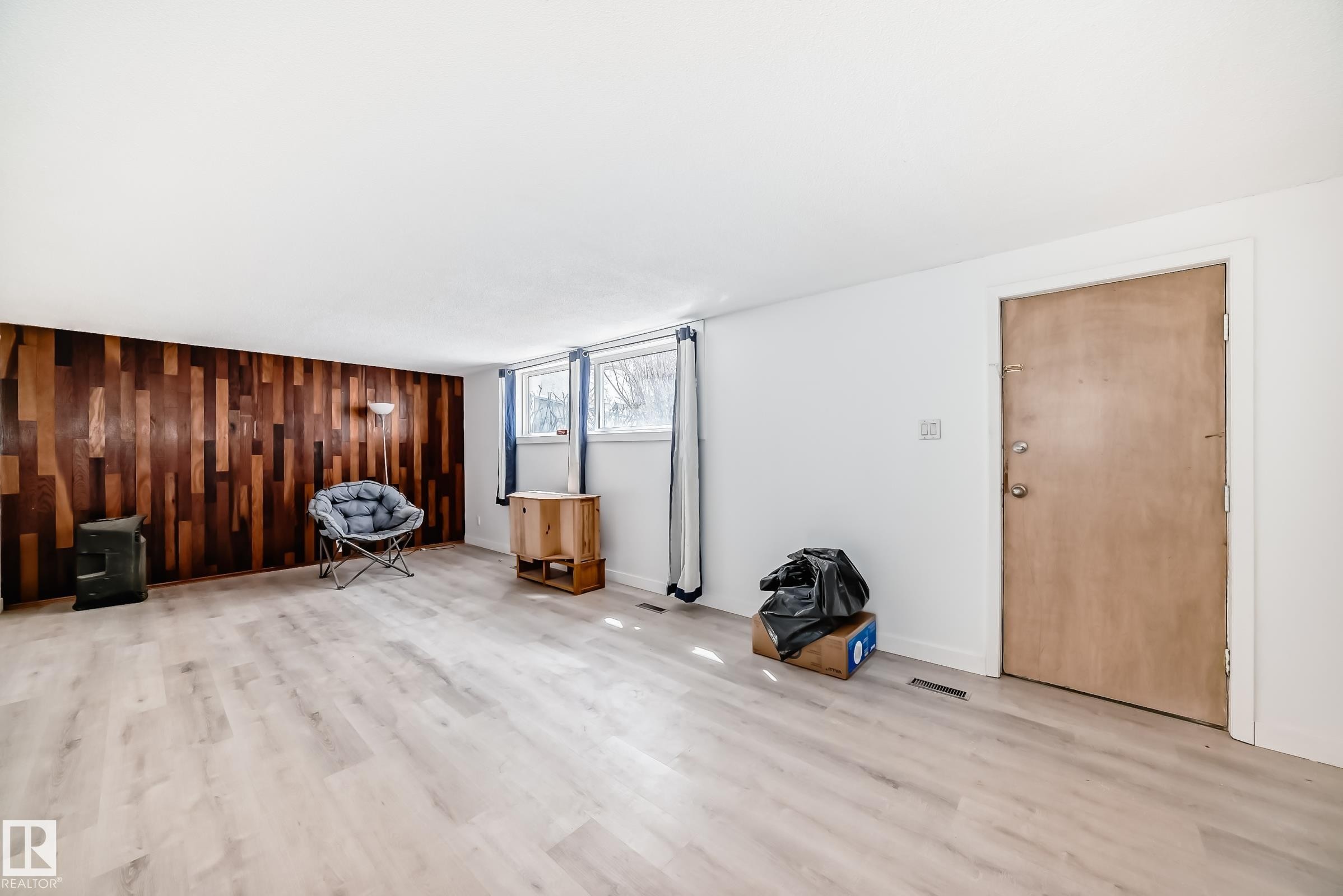 Spacious interior room featuring light wood-finish flooring, a prominent accent wall with varied wood paneling, white painted walls, and a light-colored entry door - 12825 102 Street, Edmonton, AB - Indoor Photo Showing Other Room