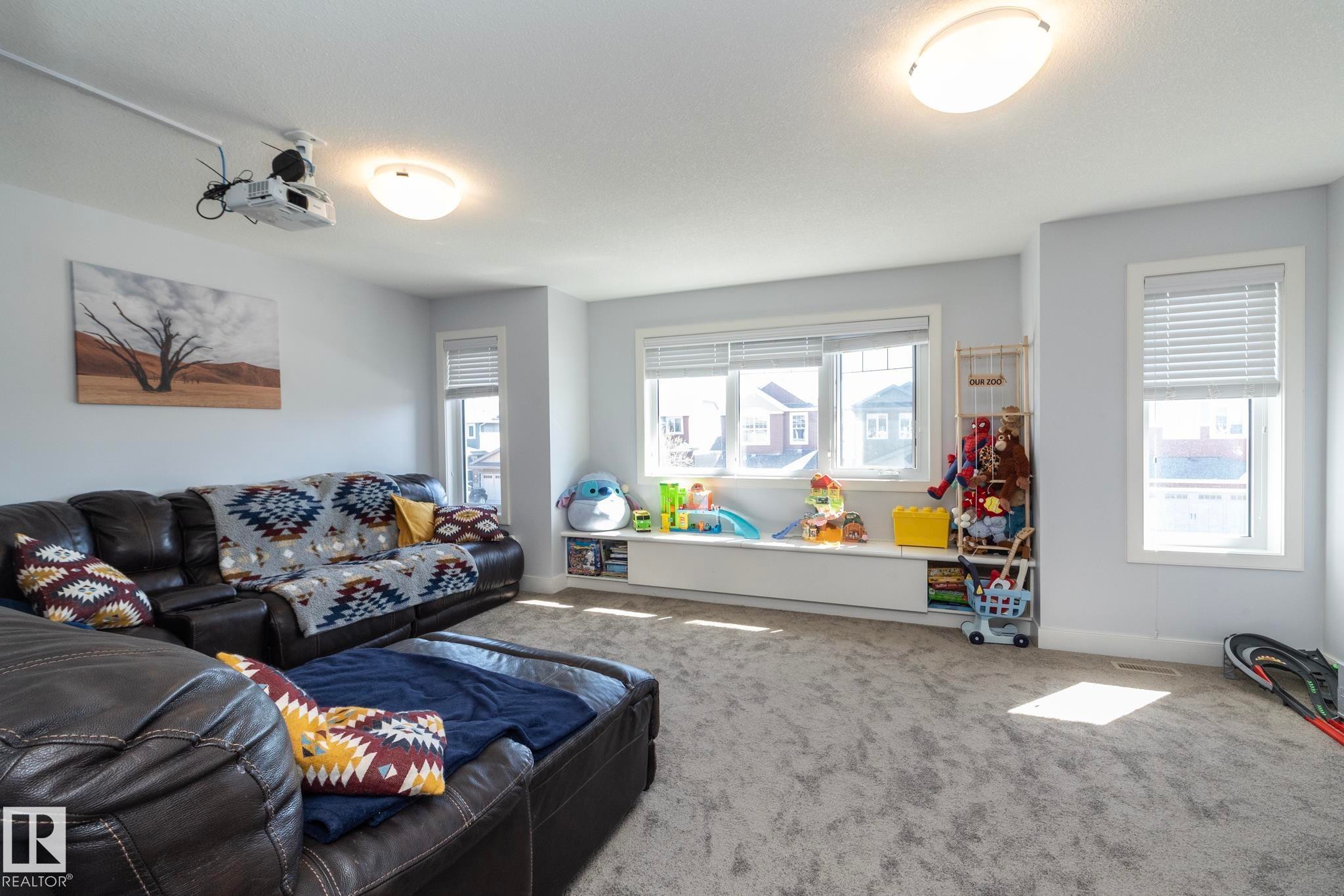 Spacious room featuring light gray wall paint, an integrated ceiling-mounted projector, multiple windows with blinds, a built-in window bench, and light gray carpeted flooring - 2016 Redtail Common, Edmonton, AB - Indoor Photo Showing Living Room