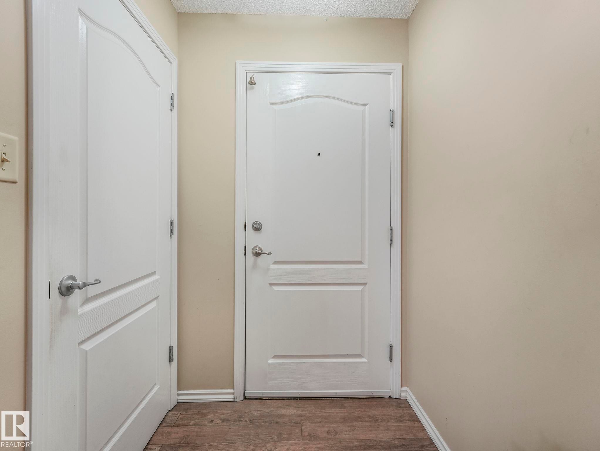 Entryway featuring a solid panel door with brushed nickel hardware, light neutral wall paint, white baseboards, and wood-finish flooring - 317 2420 108 Street, Edmonton, AB - Indoor Photo Showing Other Room