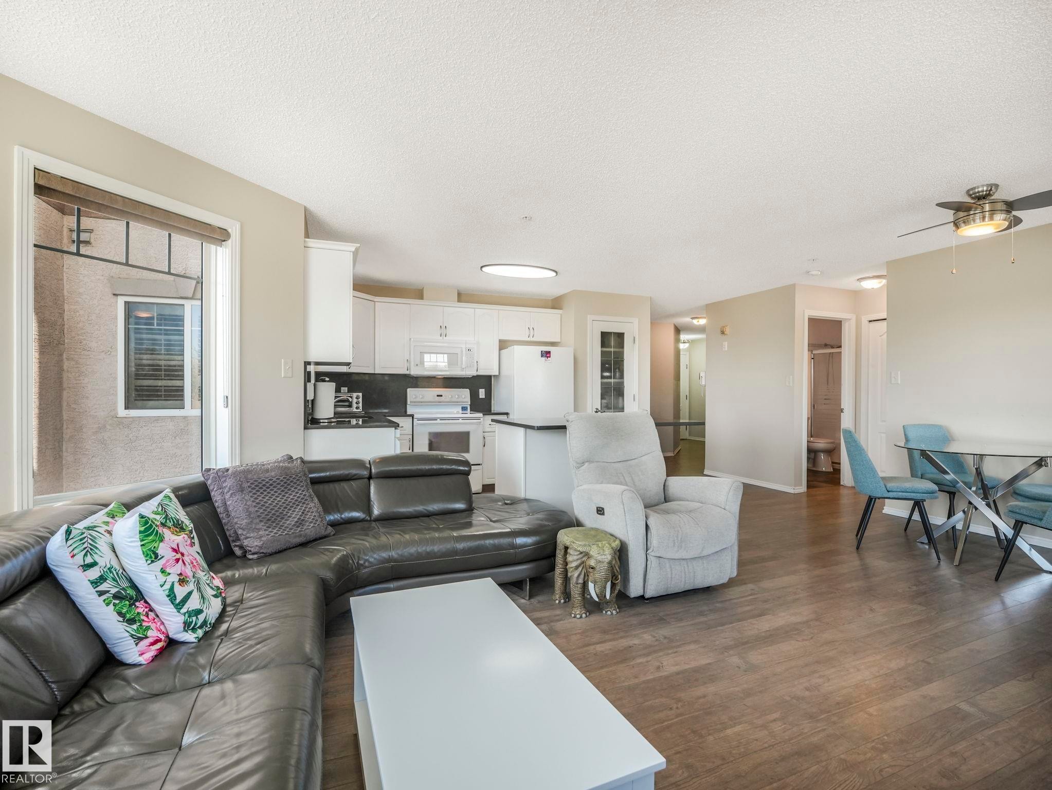 Open-concept living area featuring wood-finish flooring, a kitchen with white cabinetry and black countertops, and a dining area with a glass-top table - 317 2420 108 Street, Edmonton, AB - Indoor Photo Showing Living Room