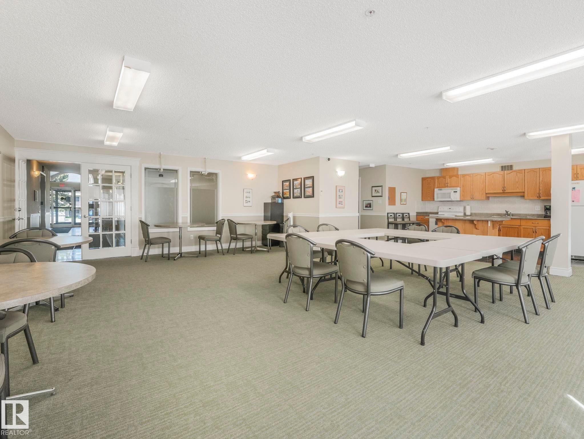 Spacious common area featuring textured carpeting, multiple tables with seating, a built-in kitchen with wood-finish cabinetry, and recessed fluorescent lighting - 317 2420 108 Street, Edmonton, AB - Indoor