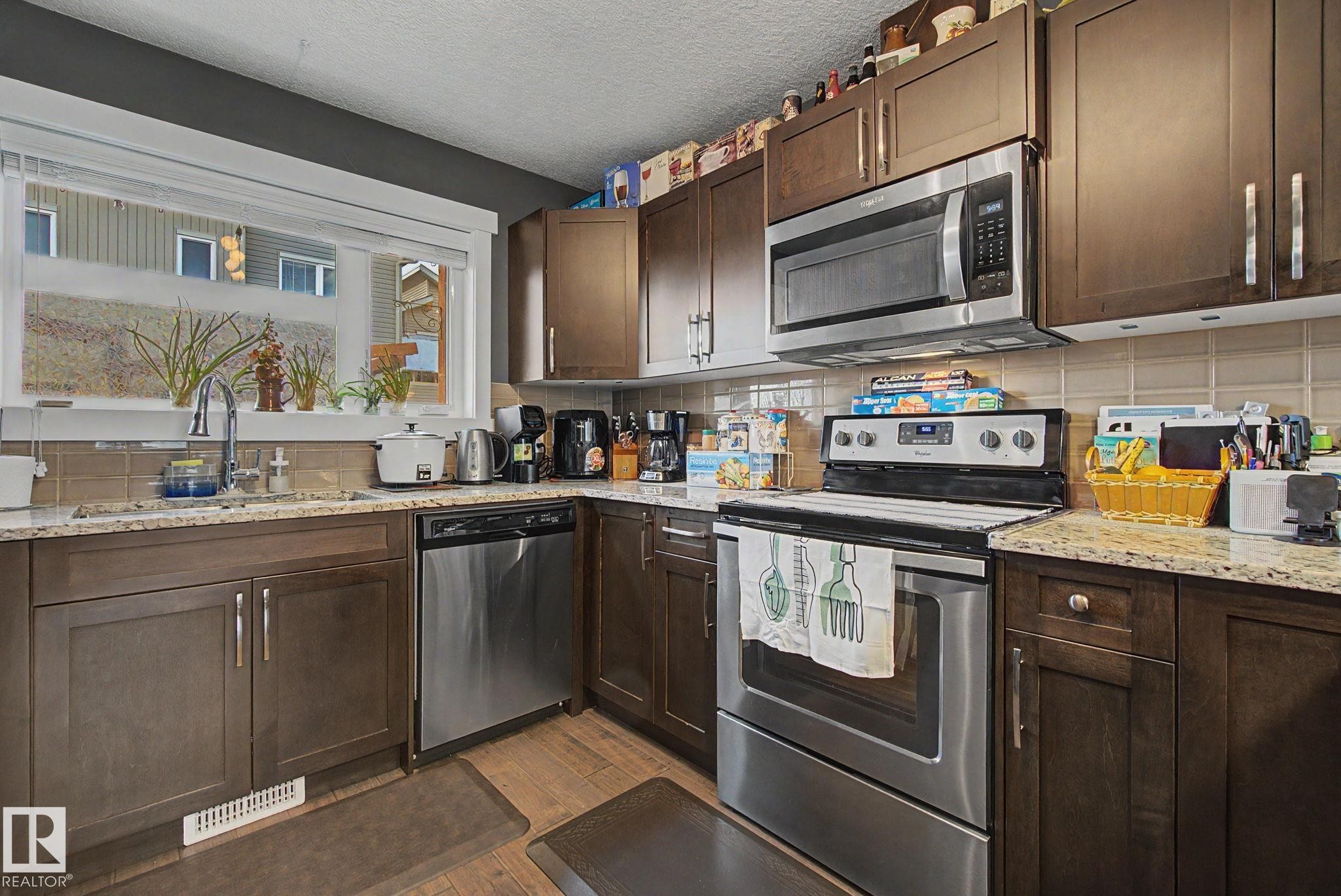 Kitchen featuring wood-finish cabinetry, granite-style countertops, stainless steel appliances, a tiled backsplash, and wood-finish flooring - 210 401 Southfork Drive, Leduc, AB - Indoor Photo Showing Kitchen