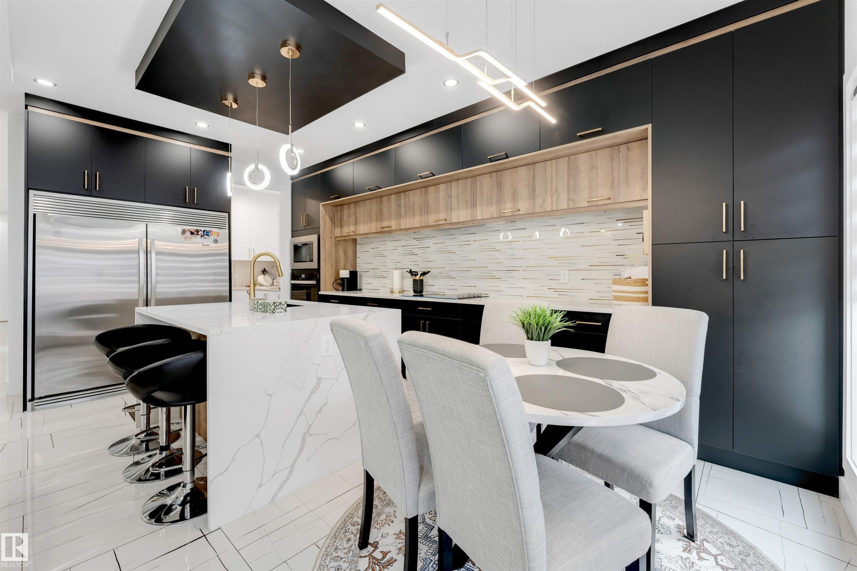 Contemporary kitchen featuring a large waterfall island with integrated seating, extensive dark cabinetry with gold-tone hardware, full-height paneled refrigeration, a light-toned wood-finish backsplash, and a gold-tone faucet - 1827 17 Avenue, Edmonton, AB - Indoor Photo Showing Dining Room