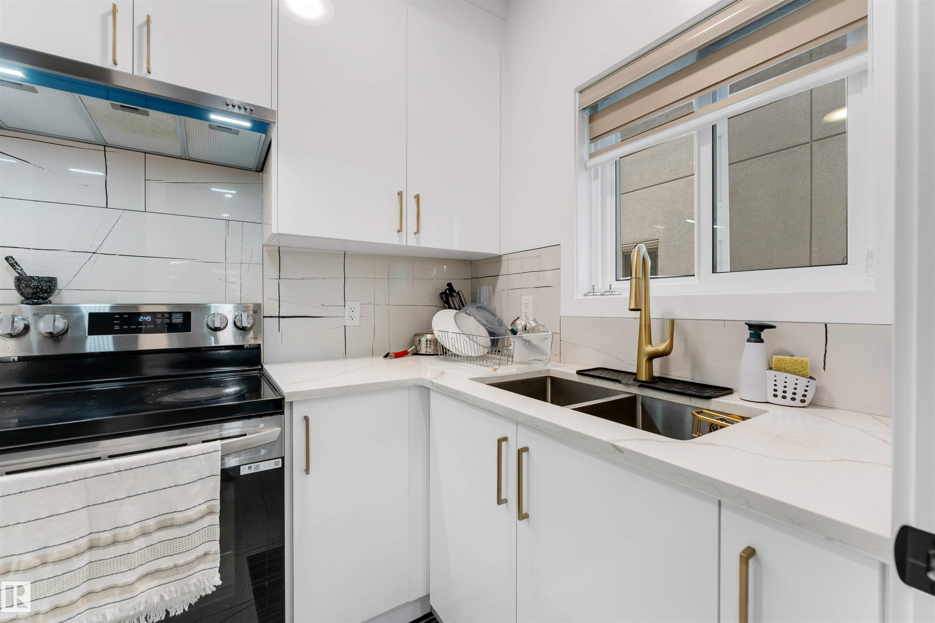 Modern kitchen featuring white cabinetry, a double basin sink with a gold-tone faucet, a stainless steel range, and light-toned countertops with a matching backsplash - 1827 17 Avenue, Edmonton, AB - Indoor Photo Showing Kitchen With Double Sink With Upgraded Kitchen