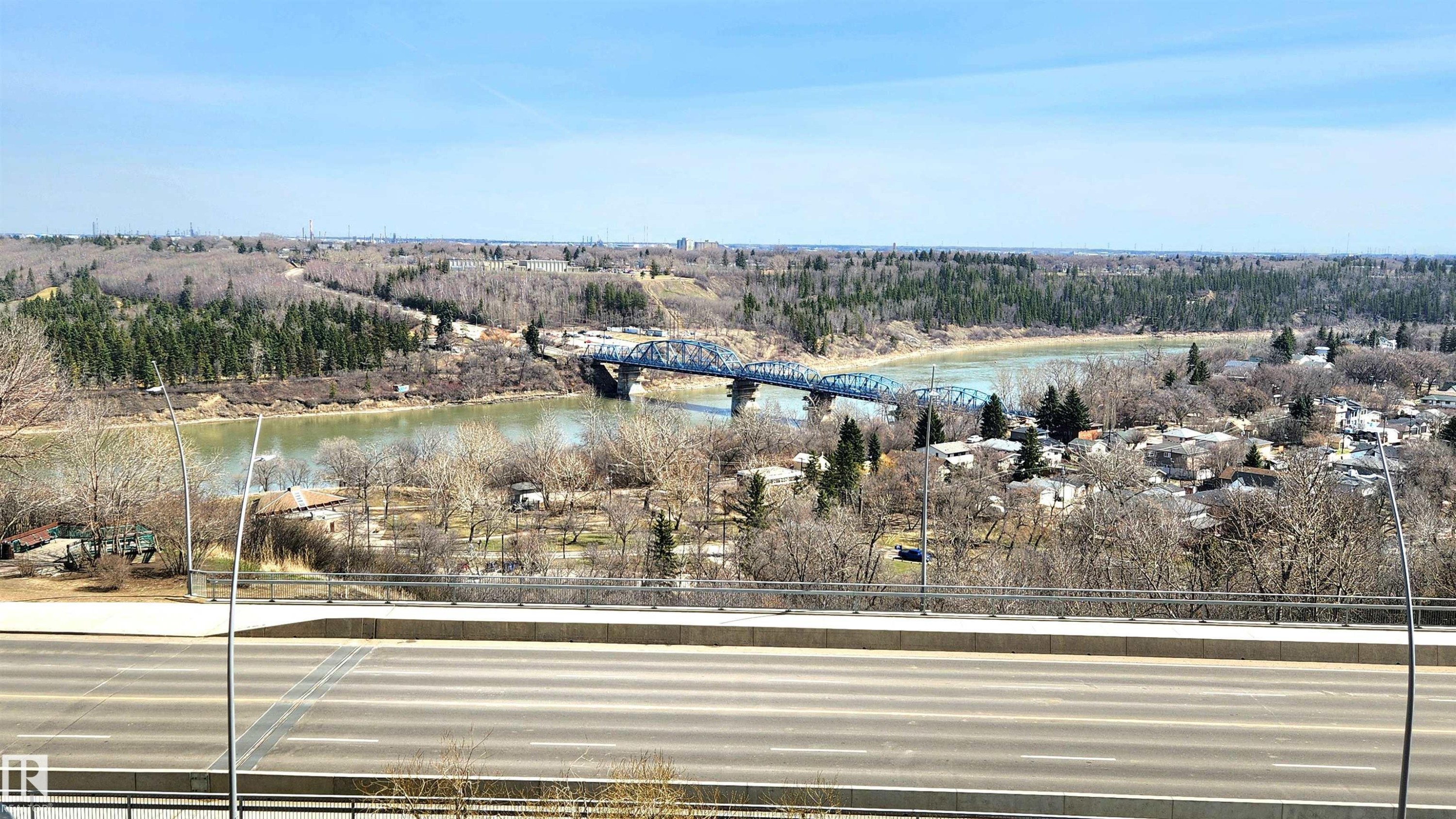 Riverfront landscape featuring a blue steel bridge, distant residential structures, and extensive wooded areas under a clear sky - 603 9028 Jasper Avenue, Edmonton, AB - Outdoor With View