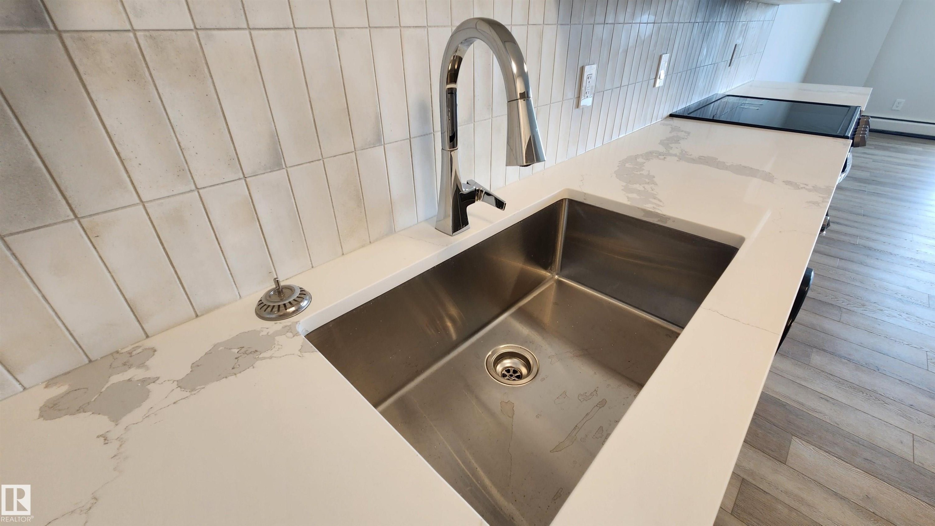 Stainless steel undermount sink with a high-arc chrome faucet, set into a white countertop with gray veining - 603 9028 Jasper Avenue, Edmonton, AB - Indoor