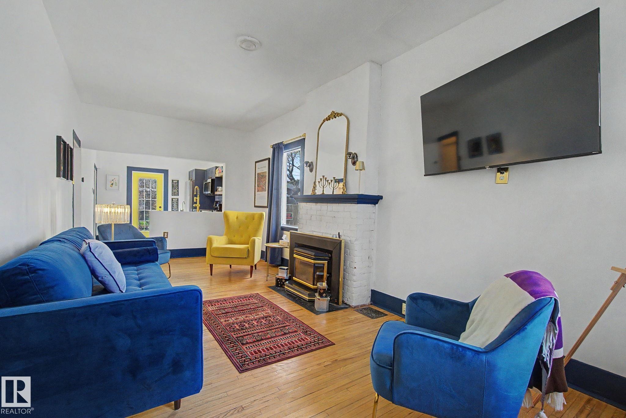 Living area featuring wood-finish flooring, a white brick fireplace with a dark mantel, a large wall-mounted display, and a half-wall partition - 11512 122 St, Edmonton, AB - Indoor Photo Showing Living Room