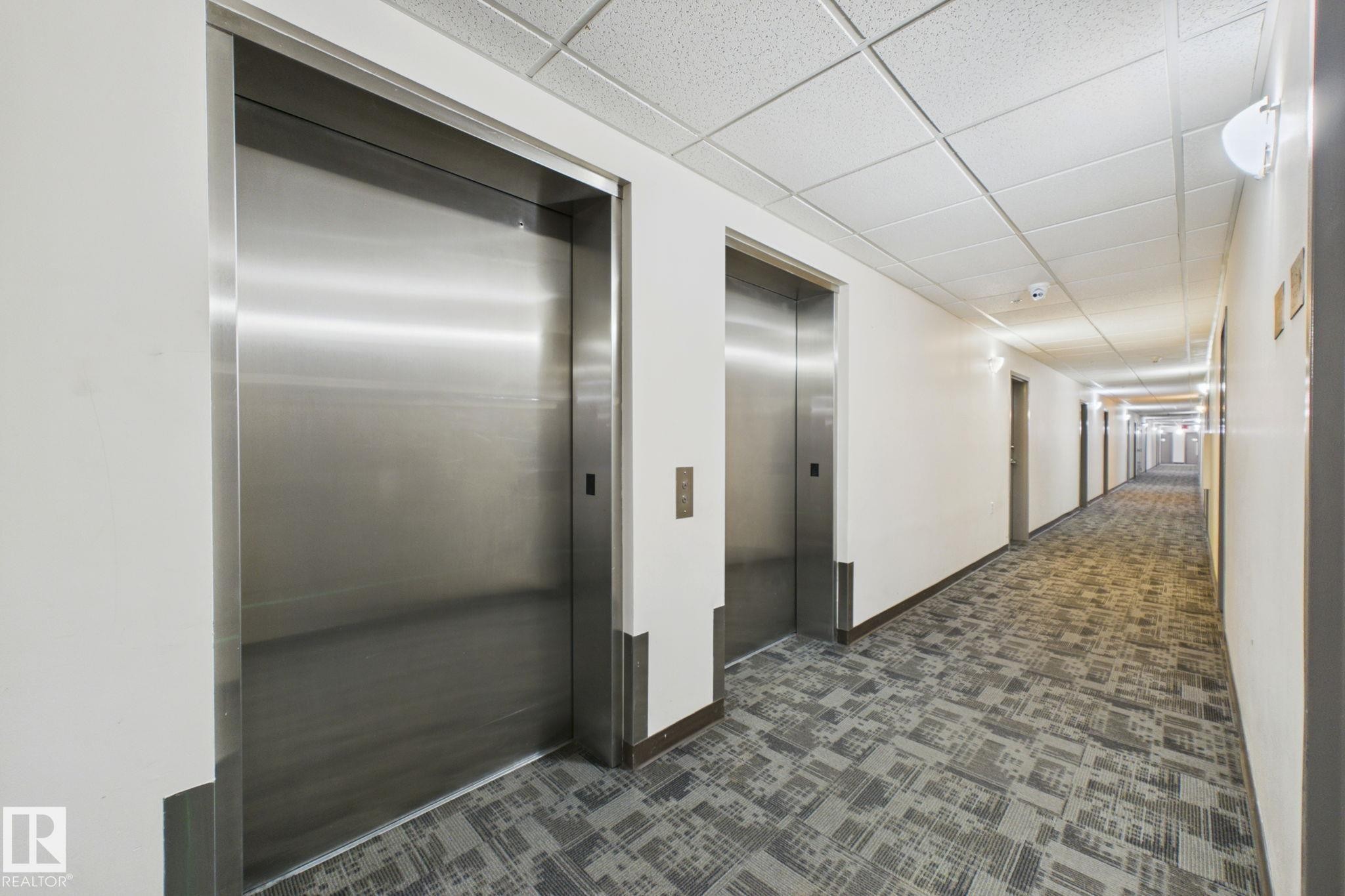 Building corridor featuring two stainless steel elevator doors, patterned carpet flooring, a drop ceiling, and recessed wall sconces - 220 80A Kelloway Crescent, Red Deer, AB - Indoor Photo Showing Other Room