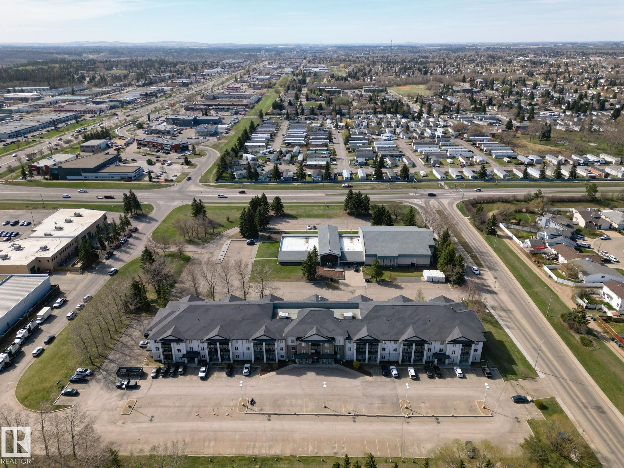 Expansive property featuring a multi-story building with a dark shingle roof and light-colored siding, a large paved parking area, and mature trees - 220 80A Kelloway Crescent, Red Deer, AB - Outdoor With View