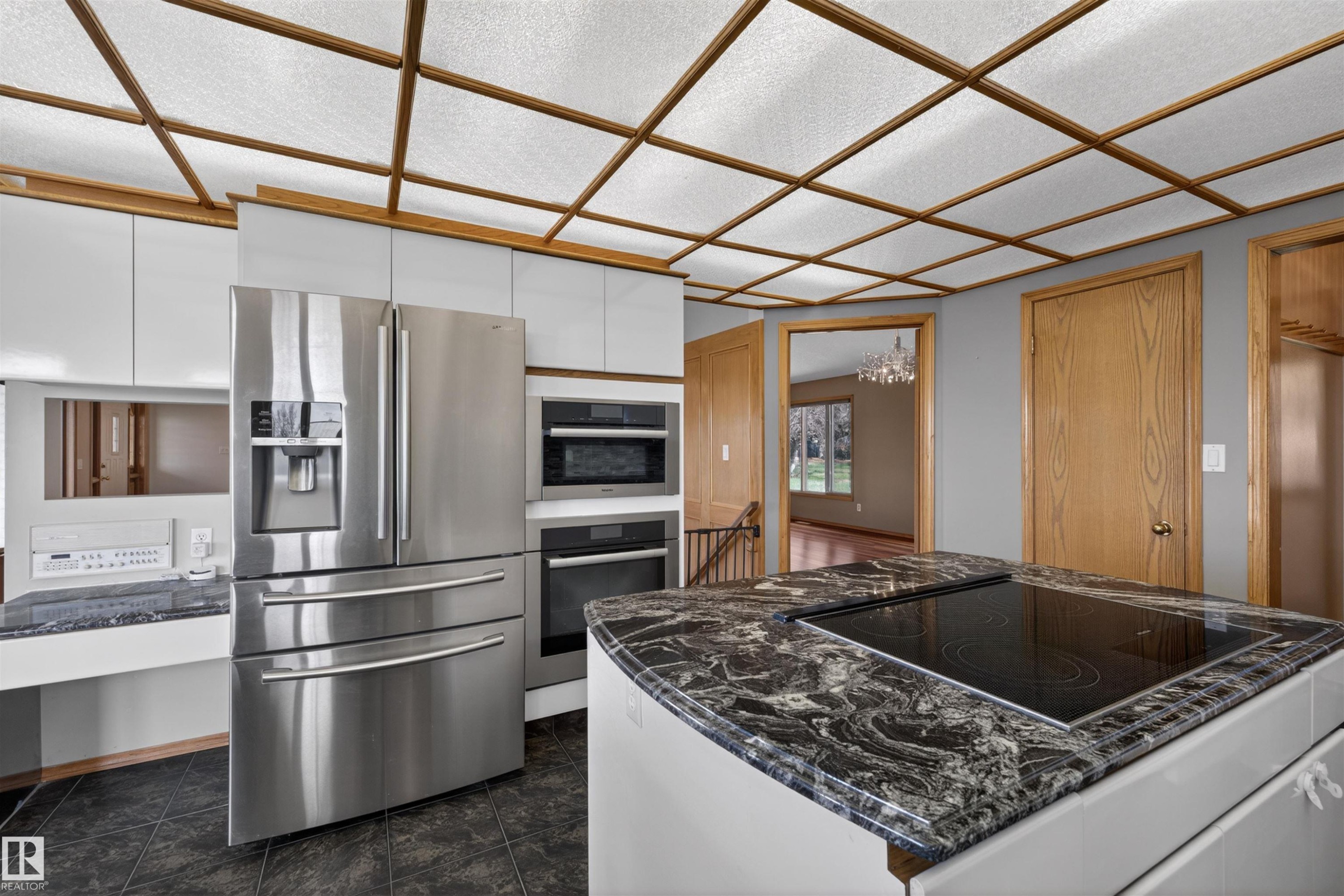 Kitchen featuring an island with a built-in cooktop and dark patterned countertop, stainless steel appliances, white cabinetry, and a grid-patterned ceiling with translucent panels - 19403 Fort Road, Edmonton, AB - Indoor Photo Showing Kitchen