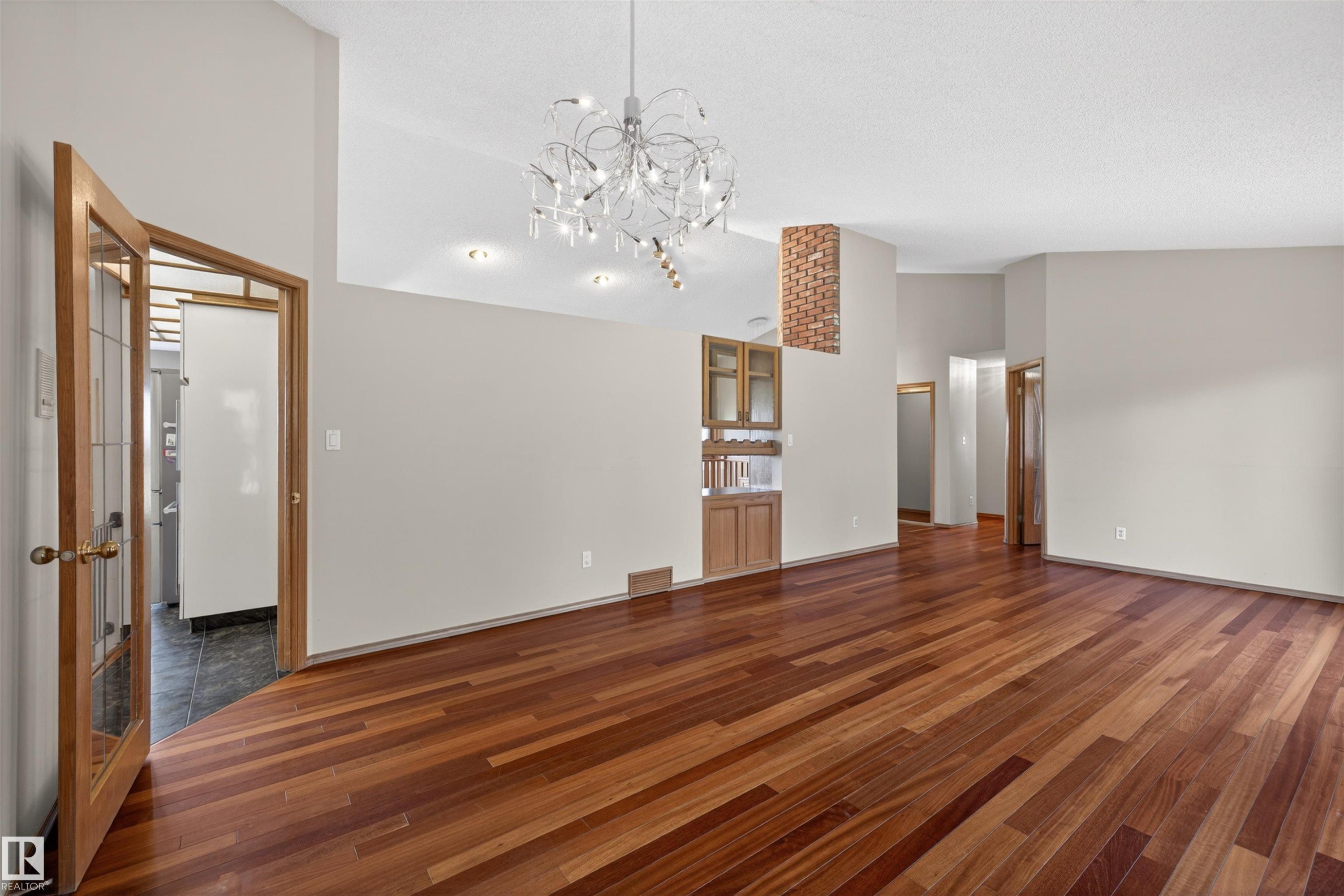 Expansive living area featuring rich wood-finish flooring, a contemporary chandelier, recessed lighting, a built-in wood cabinet with glass accents, and a distinctive exposed brick column - 19403 Fort Road, Edmonton, AB - Indoor Photo Showing Other Room