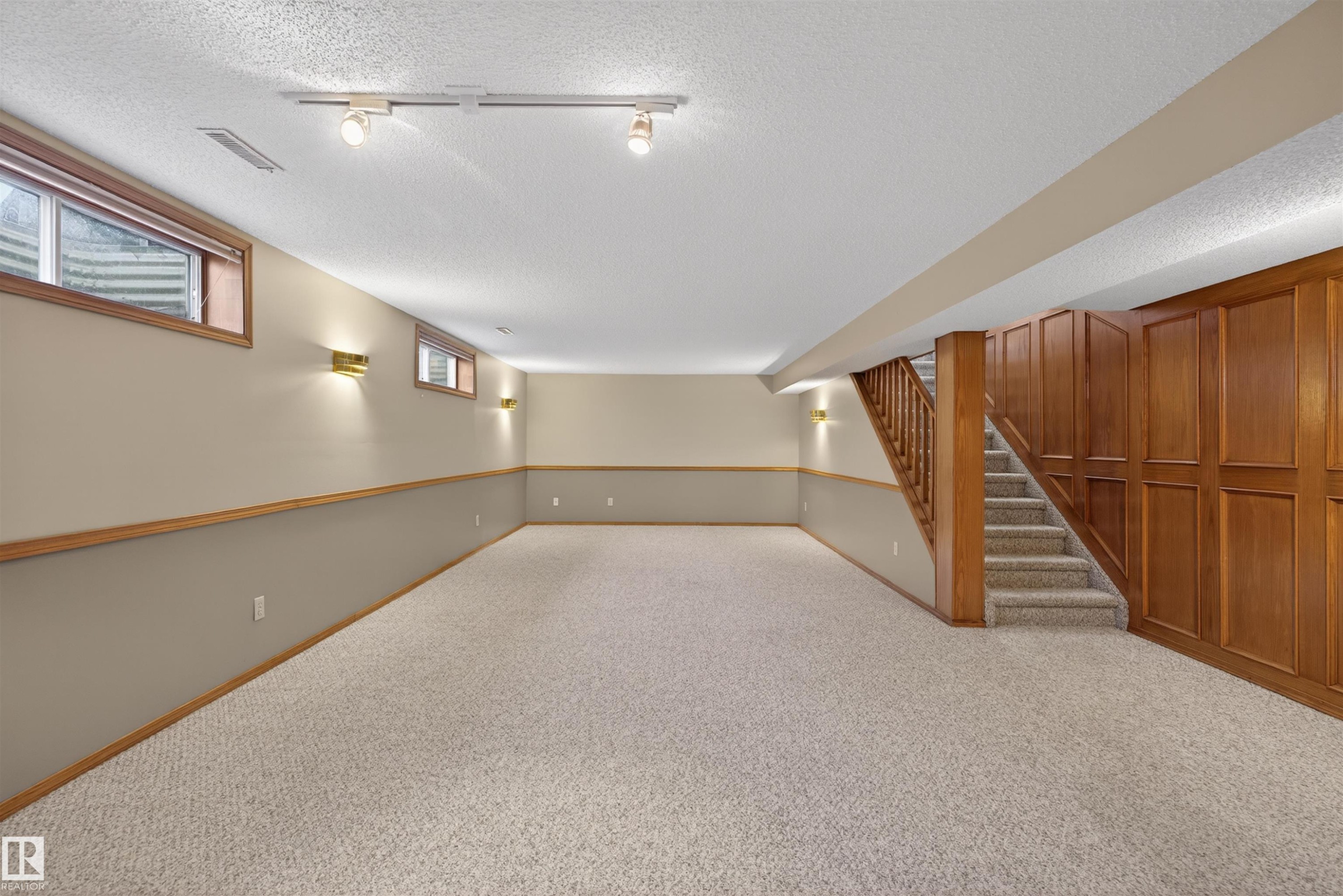 Expansive carpeted room featuring a paneled wood staircase, integrated wood cabinetry, track lighting, and multiple wall sconces - 19403 Fort Road, Edmonton, AB - Indoor Photo Showing Other Room