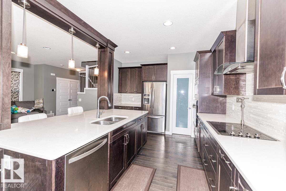 Open-concept kitchen featuring an expansive island with a stainless steel sink, dark wood-finish cabinetry, light-toned countertops, and integrated stainless steel appliances - 9748 205 Street, Edmonton, AB - Indoor Photo Showing Kitchen With Stainless Steel Kitchen With Double Sink With Upgraded Kitchen