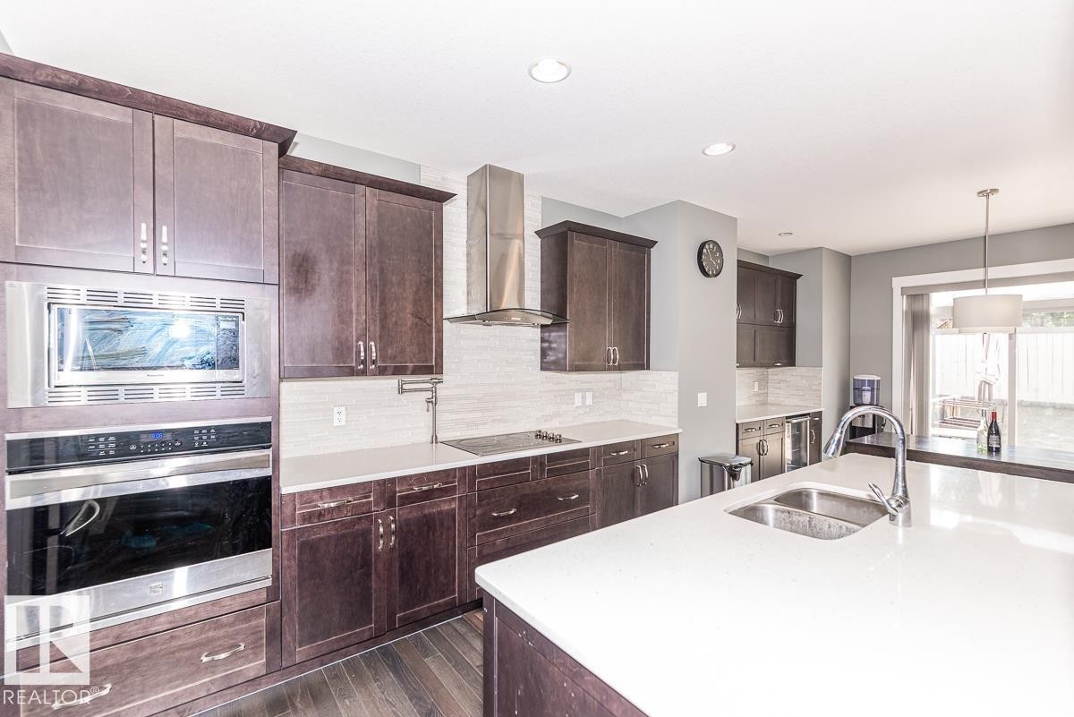 Open-concept kitchen featuring dark wood cabinetry, stainless steel appliances, a large center island with an undermount sink, light-toned countertops, and wood-finish flooring - 9748 205 Street, Edmonton, AB - Indoor Photo Showing Kitchen With Double Sink With Upgraded Kitchen