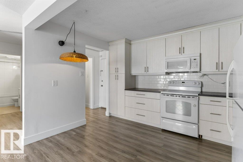 Kitchen featuring light wood-finish flooring, white cabinetry, dark countertops, a white subway tile backsplash, and white appliances - 101 8604 Gateway Boulevard, Edmonton, AB - Indoor Photo Showing Kitchen