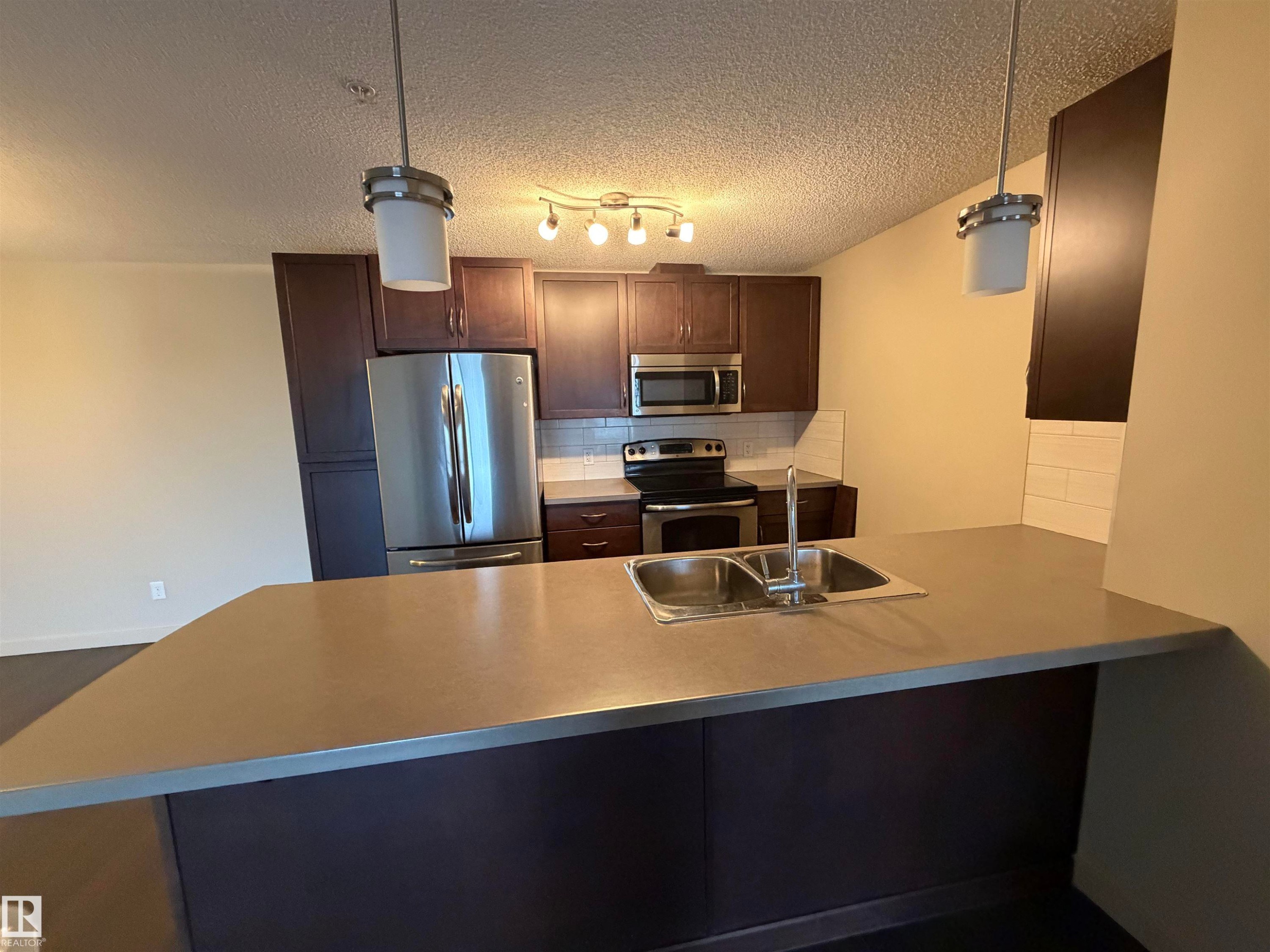 The kitchen features dark wood cabinetry, stainless steel appliances, a white subway tile backsplash, and a breakfast bar with a double basin sink - 313 6084 Stanton Drive, Edmonton, AB - Indoor Photo Showing Kitchen With Double Sink