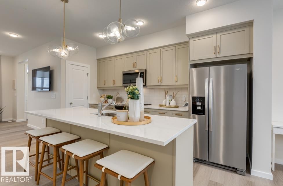 Contemporary kitchen featuring an expansive island with a light-colored countertop, overhead pendant lighting, light-toned cabinetry with gold-finish hardware, stainless steel appliances, and wood-finish flooring - 720 Southfork Drive, Leduc, AB - Indoor Photo Showing Kitchen With Upgraded Kitchen