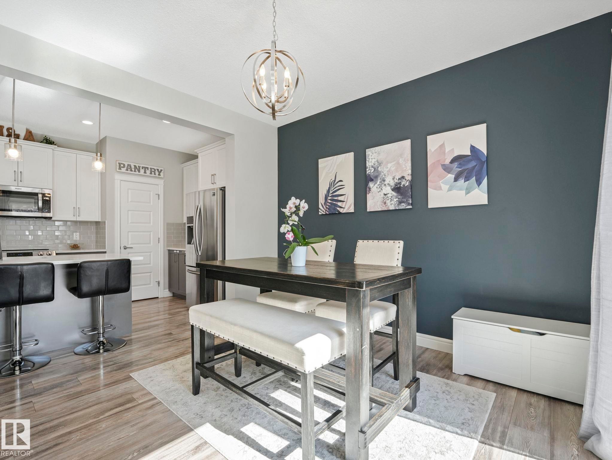 Open-concept dining area with a modern chandelier, dark accent wall, and wood-finish flooring, featuring a kitchen island with pendant lighting, white cabinetry, and stainless steel appliances - 3817 Robins Crescent, Edmonton, AB - Indoor Photo Showing Dining Room