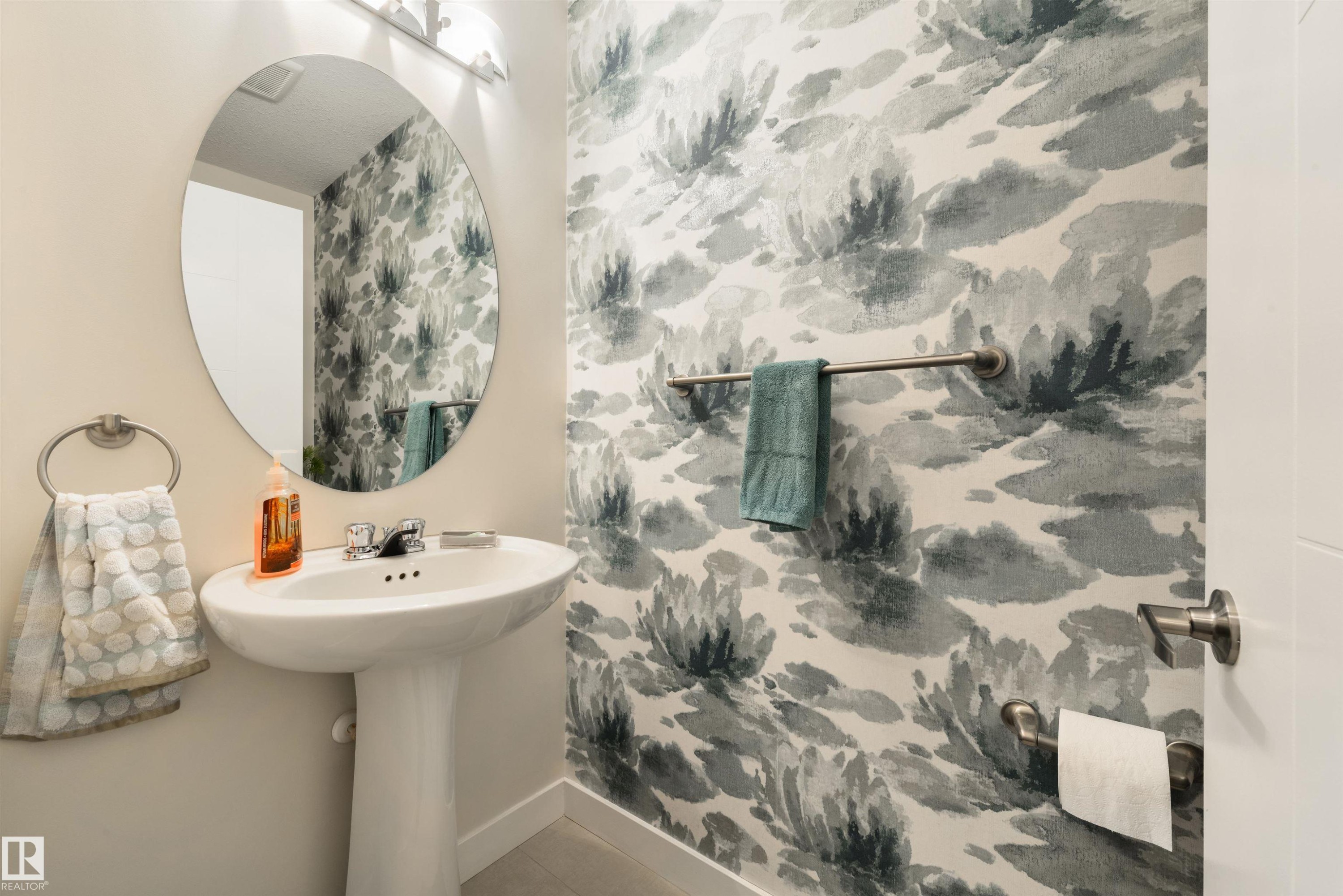 Pedestal sink with chrome faucet, oval mirror, wall-mounted lighting, and an accent wall featuring patterned wallpaper - 4914 47 Street, Redwater, AB - Indoor Photo Showing Bathroom