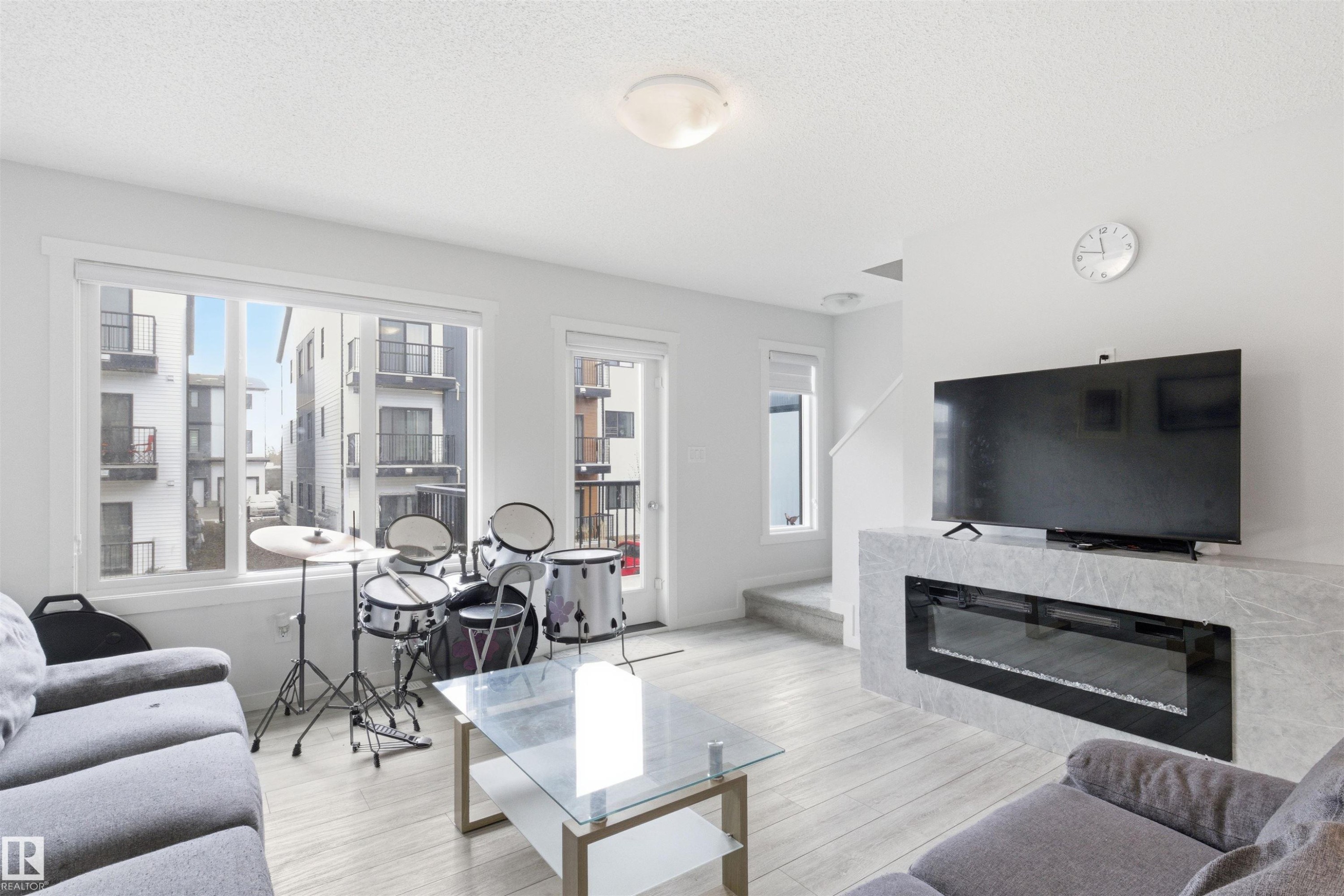 Living area featuring wood-finish flooring, a wall-mounted electric fireplace with a stone-look surround, and abundant natural light from large windows and a glass-paneled door - 9 850 Secord Boulevard, Edmonton, AB - Indoor Photo Showing Living Room With Fireplace
