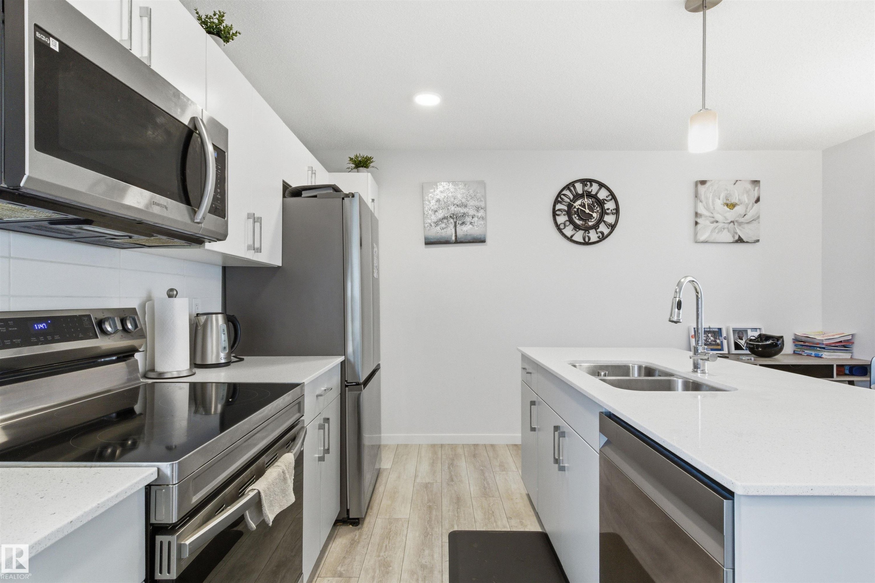 Modern kitchen featuring a central island with double basin sink, stainless steel appliances, white cabinetry, wood-finish flooring, and recessed lighting - 9 850 Secord Boulevard, Edmonton, AB - Indoor Photo Showing Kitchen With Double Sink