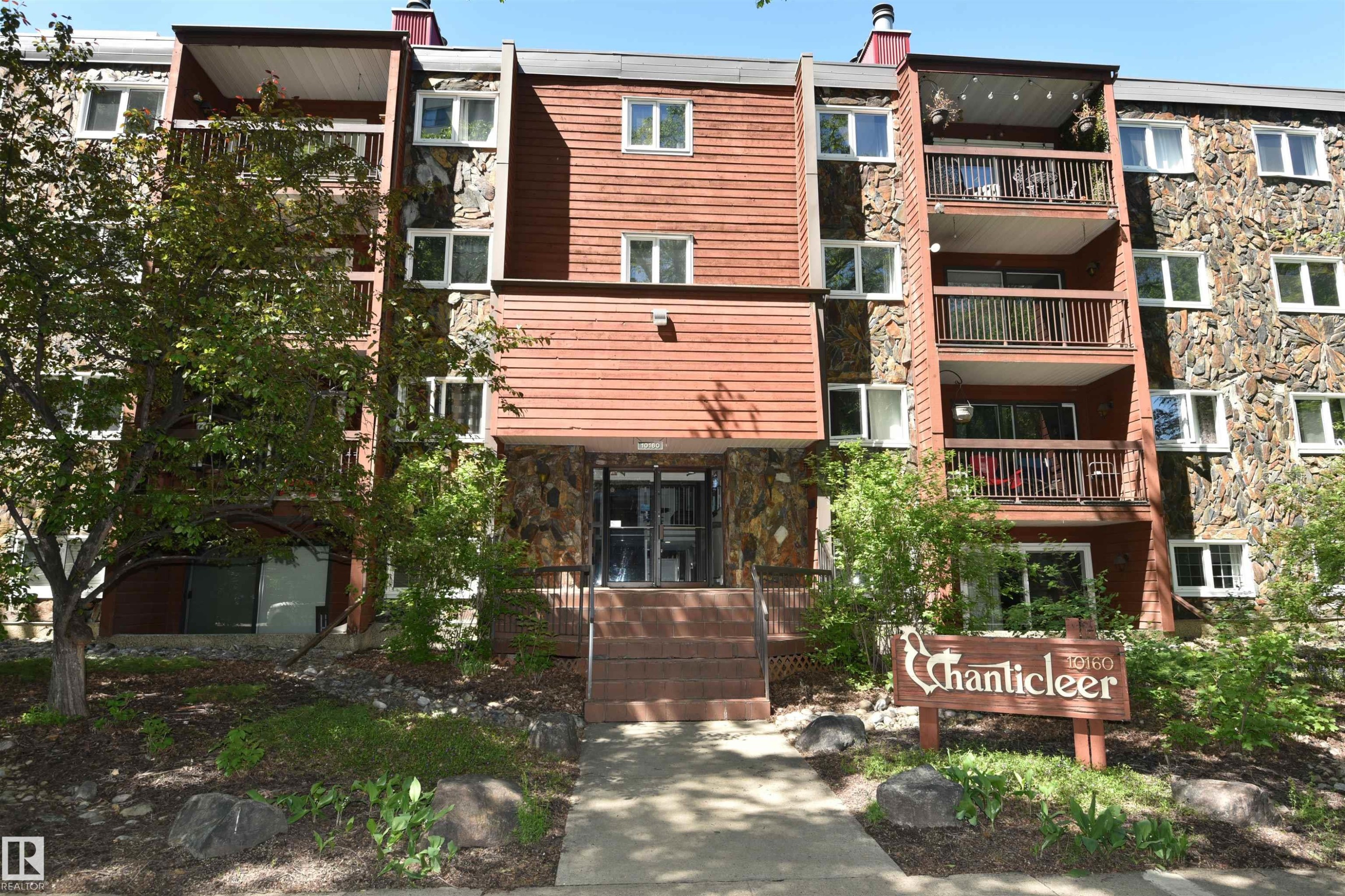 Multi-story residential building featuring a stone and wood-panel exterior, multiple balconies with dark railings, and a prominent entrance with steps - 21 10160 119 Street Nw, Edmonton, AB