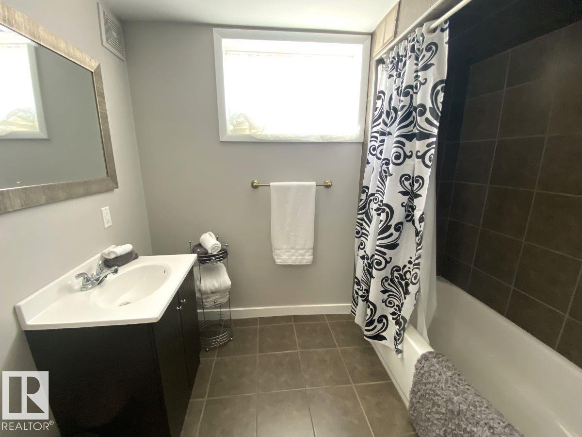 Bathroom featuring a white vanity with integrated sink, a framed mirror, a bathtub with dark-tiled surround, a window, and tile flooring - 8239 93A Avenue, Edmonton, AB - Indoor Photo Showing Bathroom
