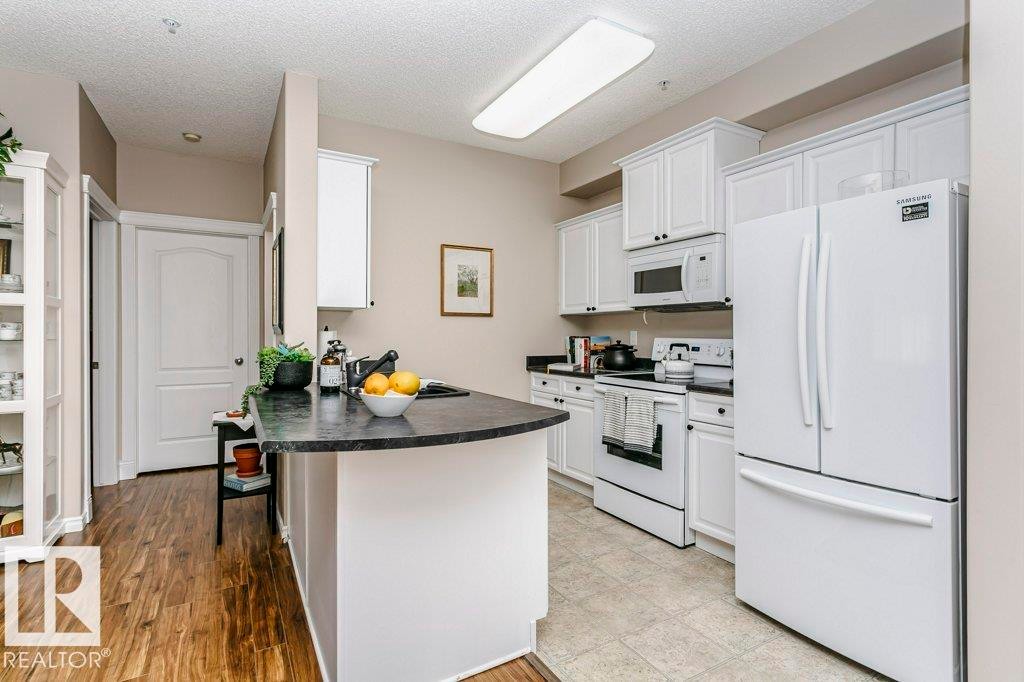 Open-concept kitchen featuring white cabinetry, contrasting dark countertops, and a white appliance suite - 323 16807 100 Avenue, Edmonton, AB - Indoor Photo Showing Kitchen