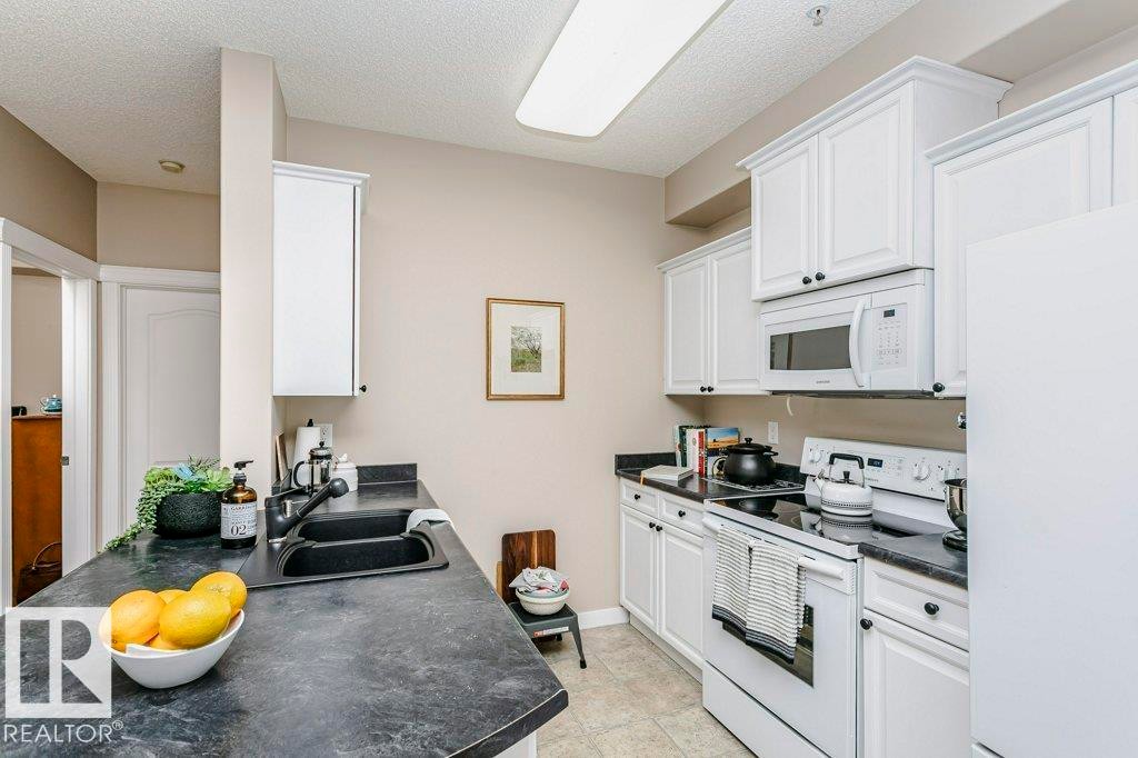Bright kitchen featuring white cabinetry, dark countertops, and a white appliance suite including an oven with an electric cooktop and an over-the-range microwave - 323 16807 100 Avenue, Edmonton, AB - Indoor Photo Showing Kitchen With Double Sink
