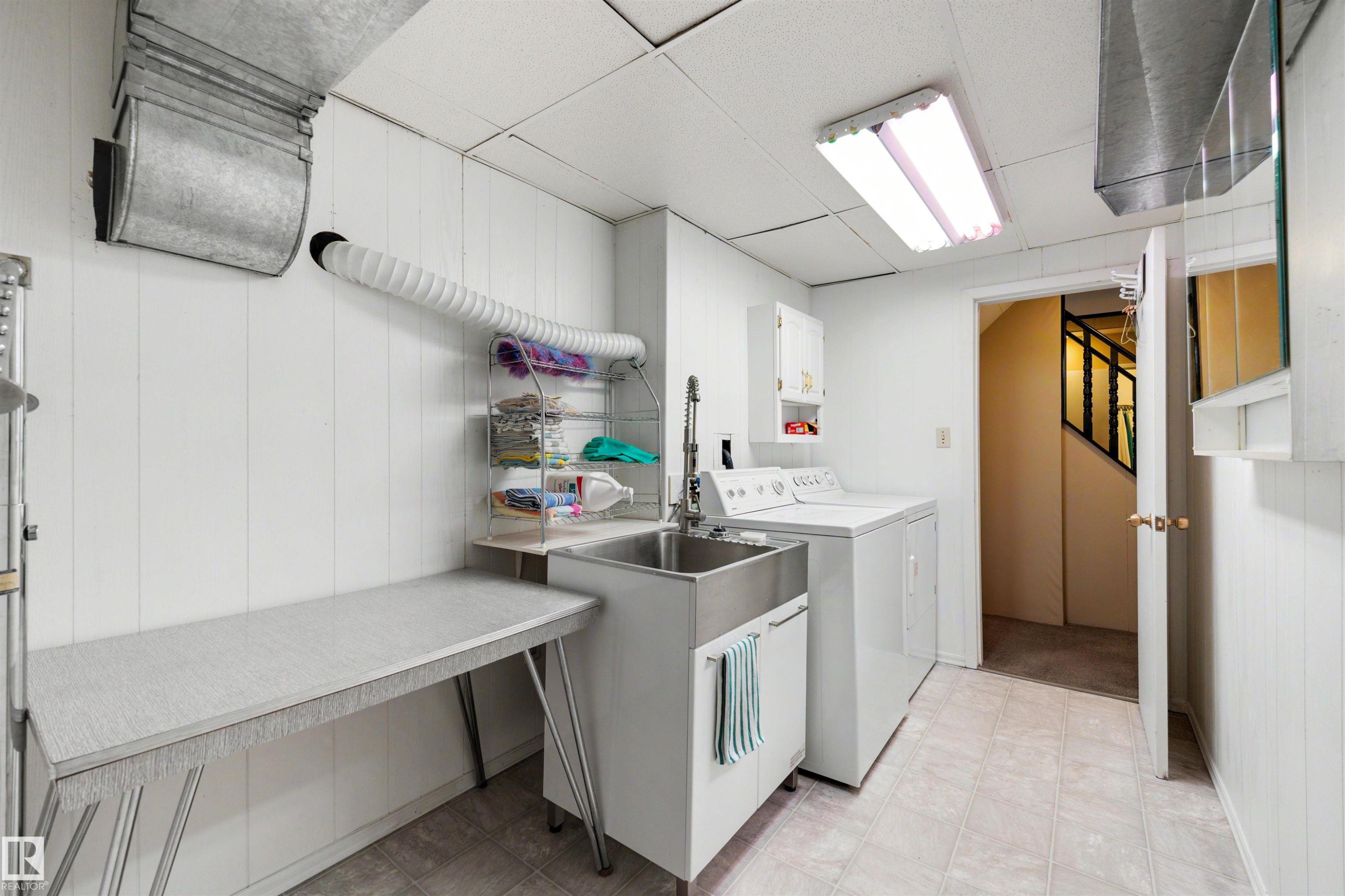 Utility room featuring a stainless steel sink, white paneled walls, a large folding surface, and tile flooring - 9602 85 Street, Edmonton, AB