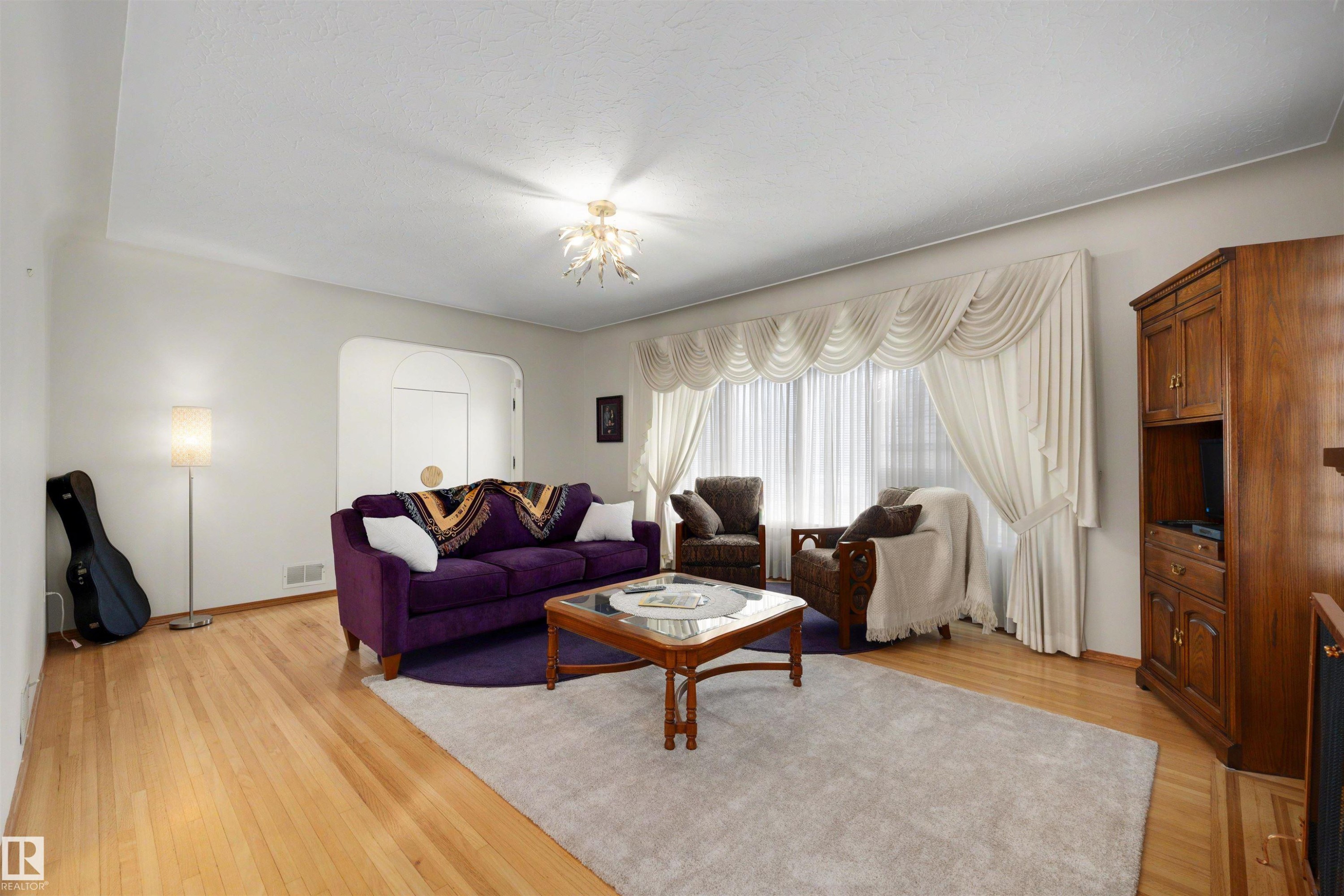 Living area featuring light wood-finish flooring, an arched doorway, a decorative ceiling fixture, and a large window with draped window treatments - 9602 85 Street, Edmonton, AB